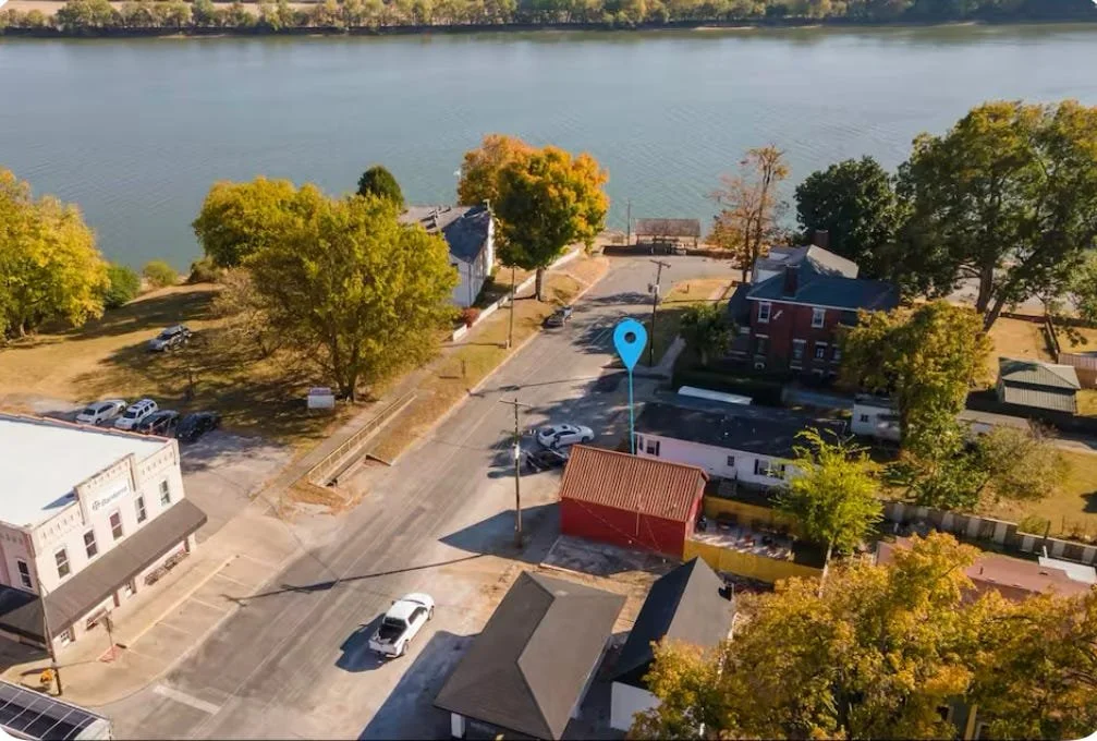 Aerial view of a lakeside town street with trees showing fall colors, parked cars, buildings, and a body of water in the background.