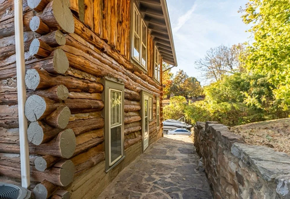 Side view of a rustic log cabin with multiple windows, a stone pathway, and a stone retaining wall, surrounded by green trees and clear sky.