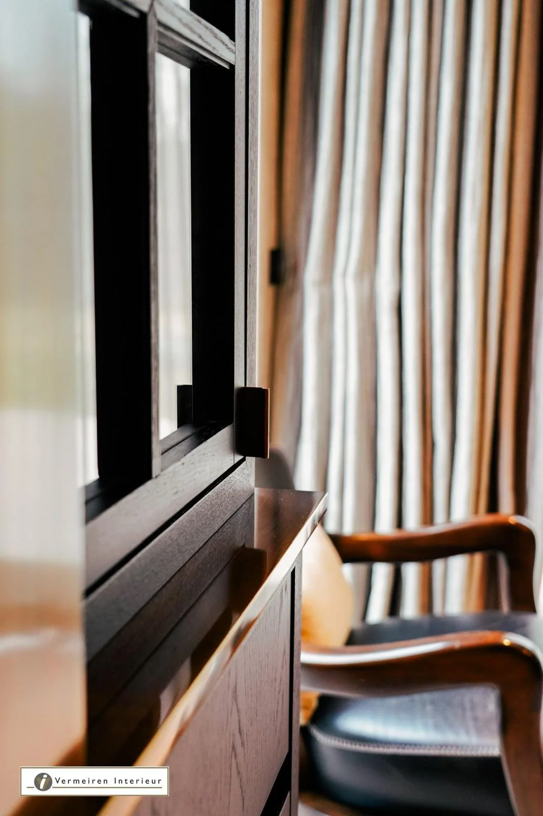 Close-up of a dark wooden cabinet or furniture piece with a window or mirror on the door and a black handle. In the background, there are beige and brown striped curtains and a wooden chair with black seating.