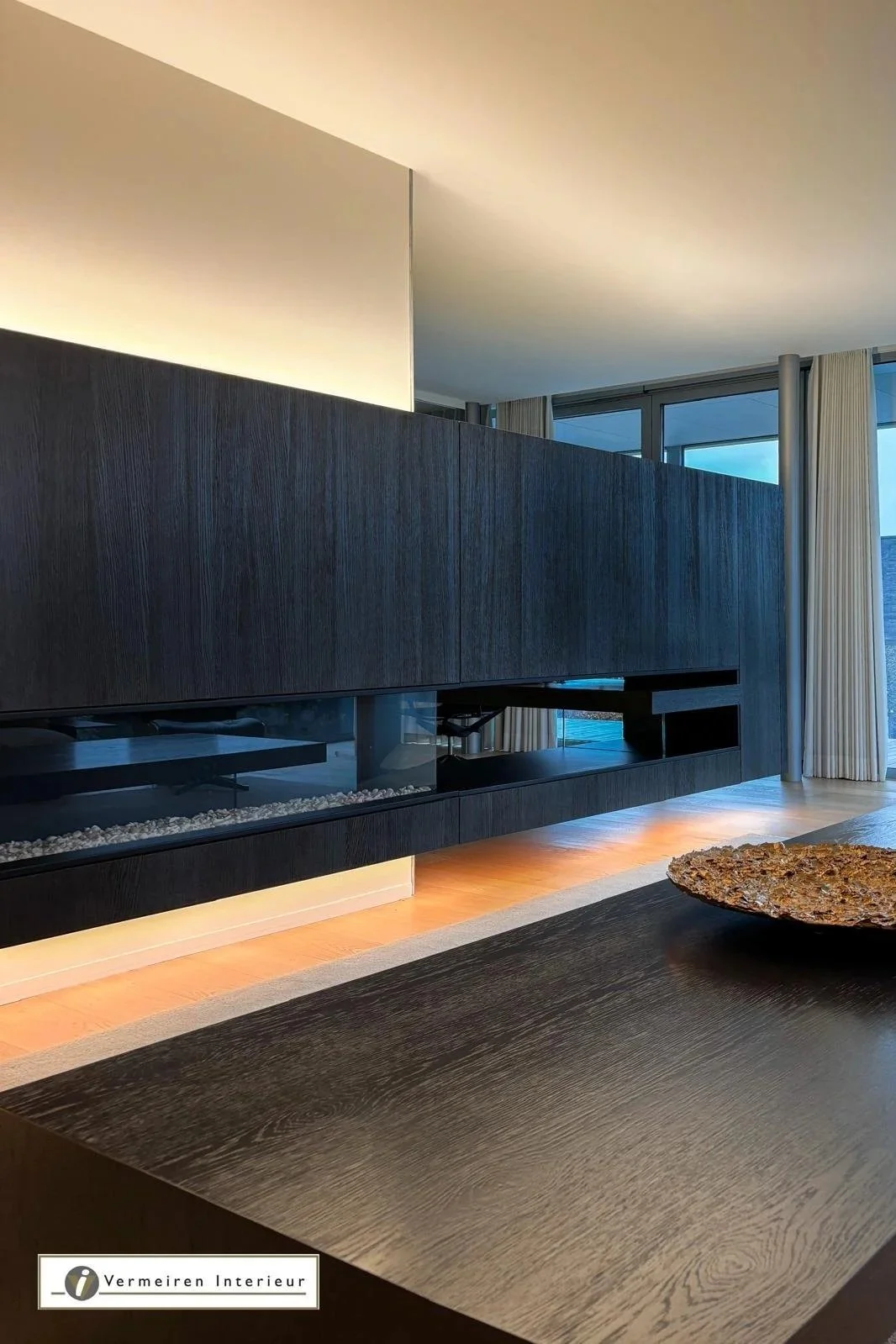 Modern living room with a black wooden fireplace on a dark wood wall, large windows with white curtains, and a wooden coffee table with a rustic bowl on it.