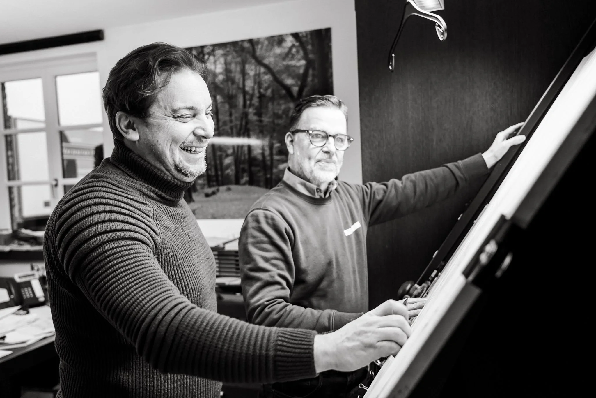 Two men playing piano in an office, one smiling and the other looking at sheet music.