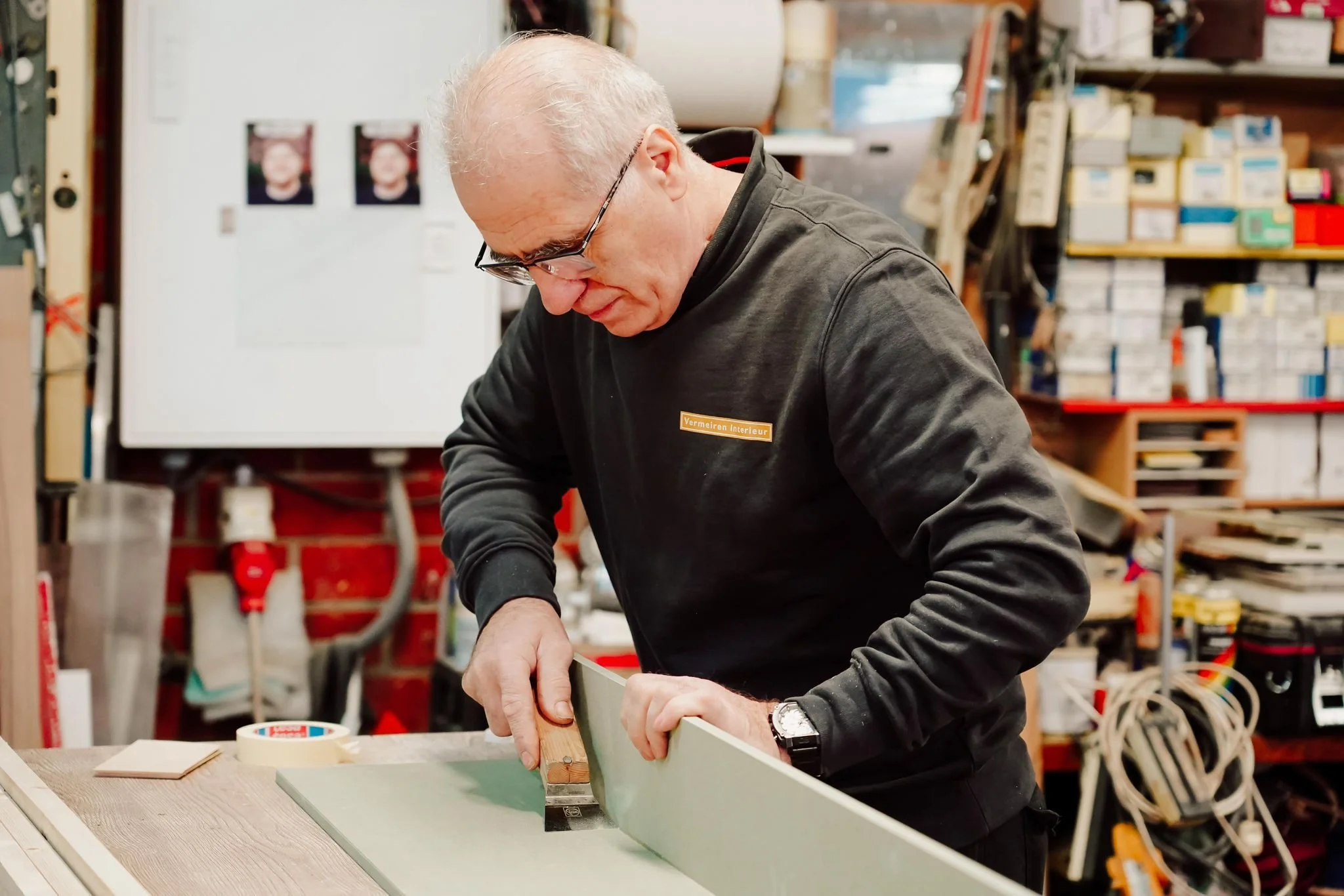 An older man with glasses working on a woodworking project in a workshop filled with shelves and tools.