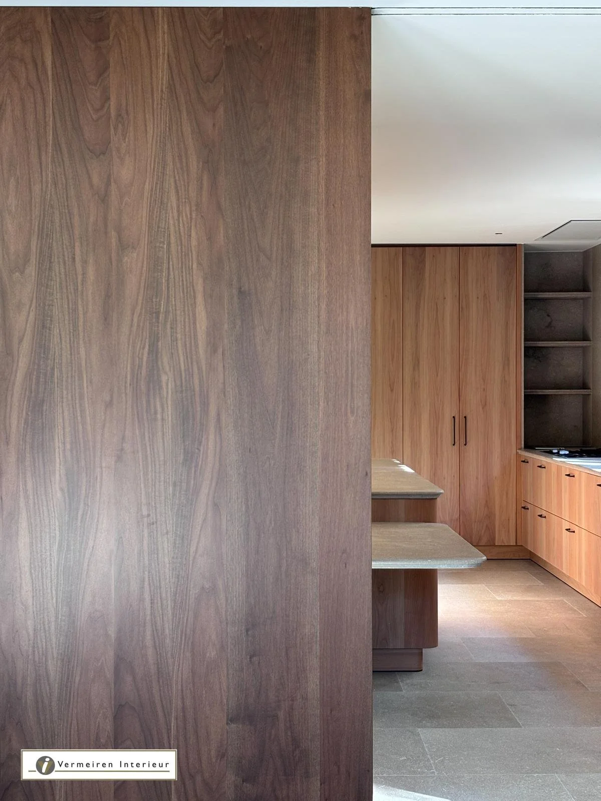 Modern kitchen with wooden cabinets and shelves, beige countertops, and a large wooden partition or wall. The floor is tiled in gray.