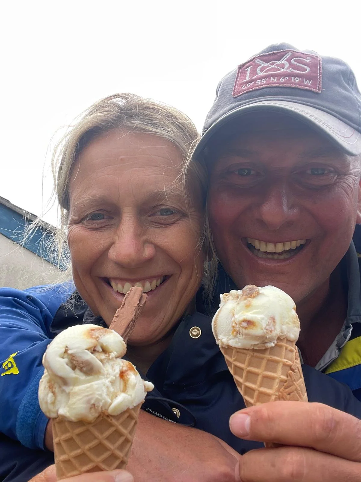 Smiling woman and man holding ice cream cones, the woman has a chocolate-covered cone, both are outdoors, overcast sky.