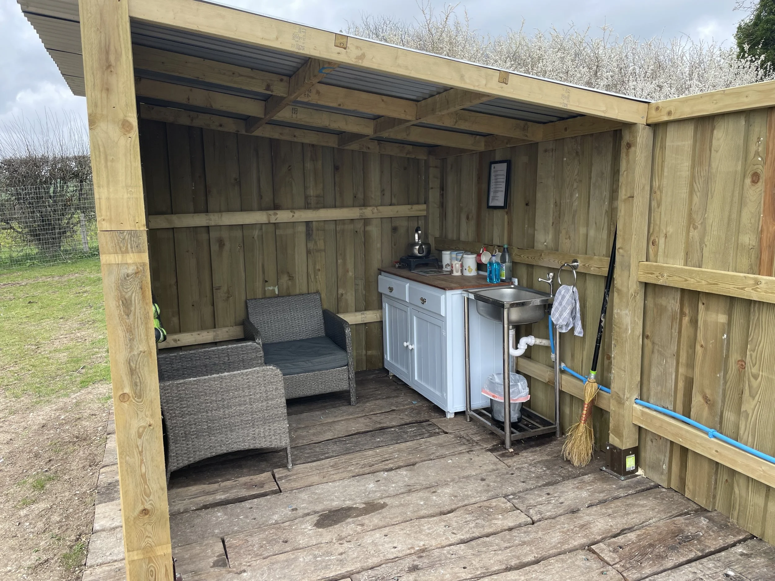 Wooden outdoor shelter with a small kitchen area, including a sink, refrigerator, and countertop, with some chairs and miscellaneous items inside.