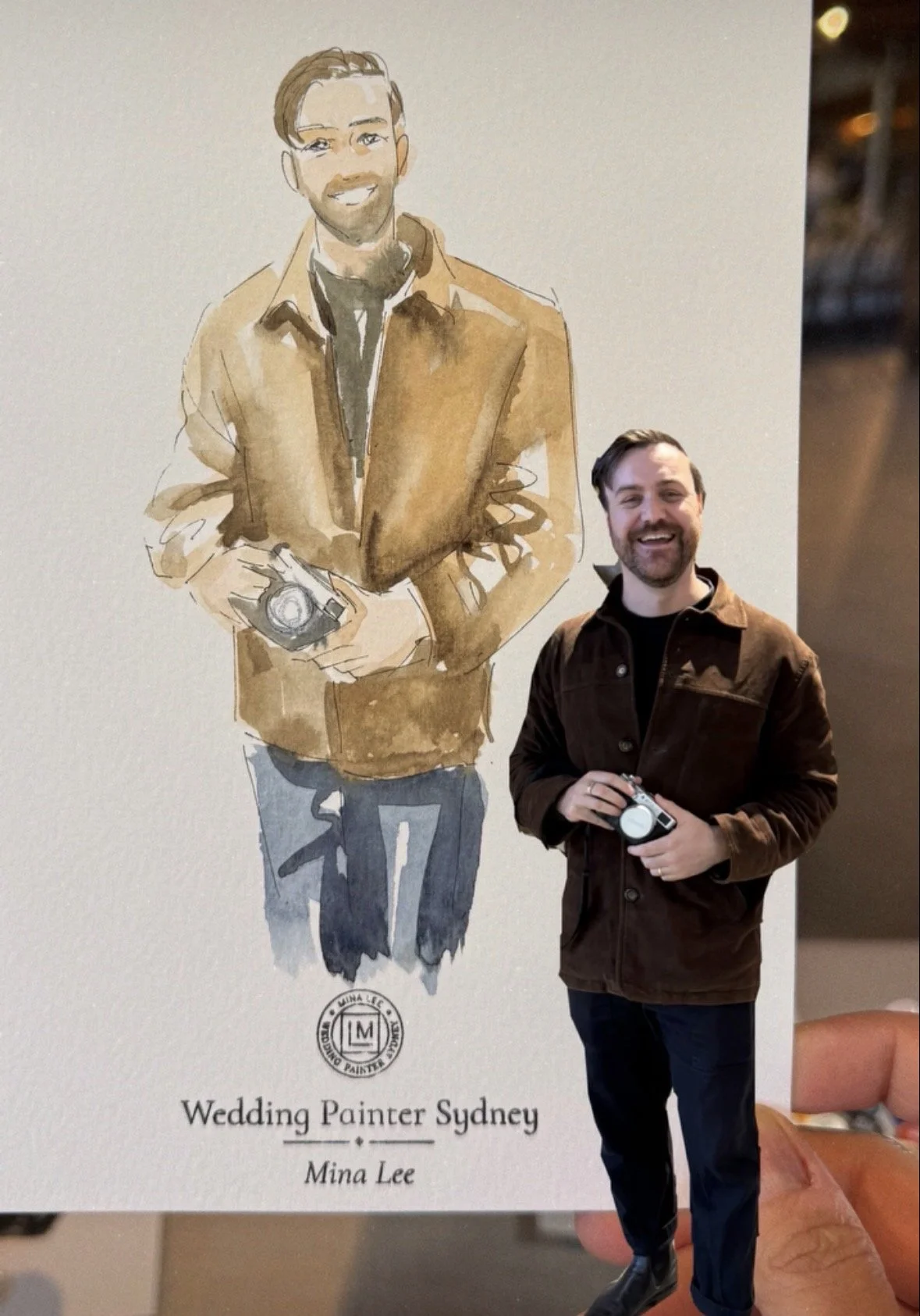 A person holding a photo of himself with a graphic illustration of a man holding a camera, with the text 'Wedding Painter Sydney' and the name 'Mina Lee' below.