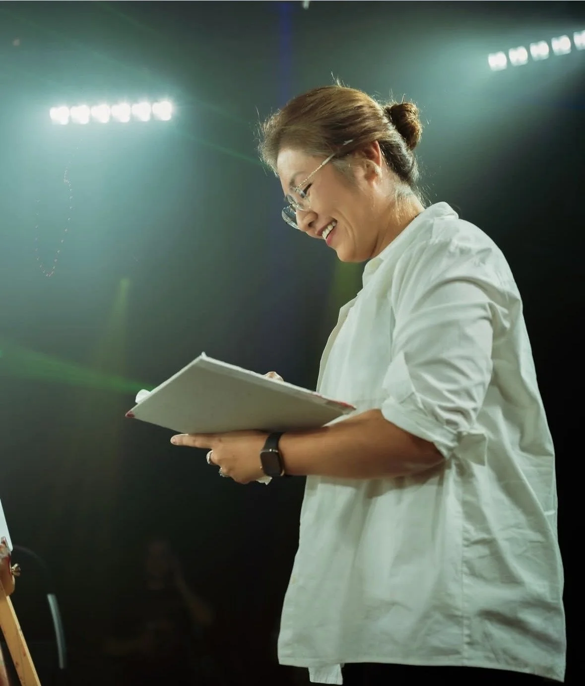 A woman with glasses, smiling and holding a script, on stage with bright lights overhead.
