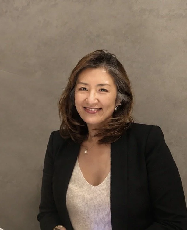 Portrait of a smiling woman with shoulder-length wavy hair, wearing a black blazer over a white top, standing against a plain gray background.