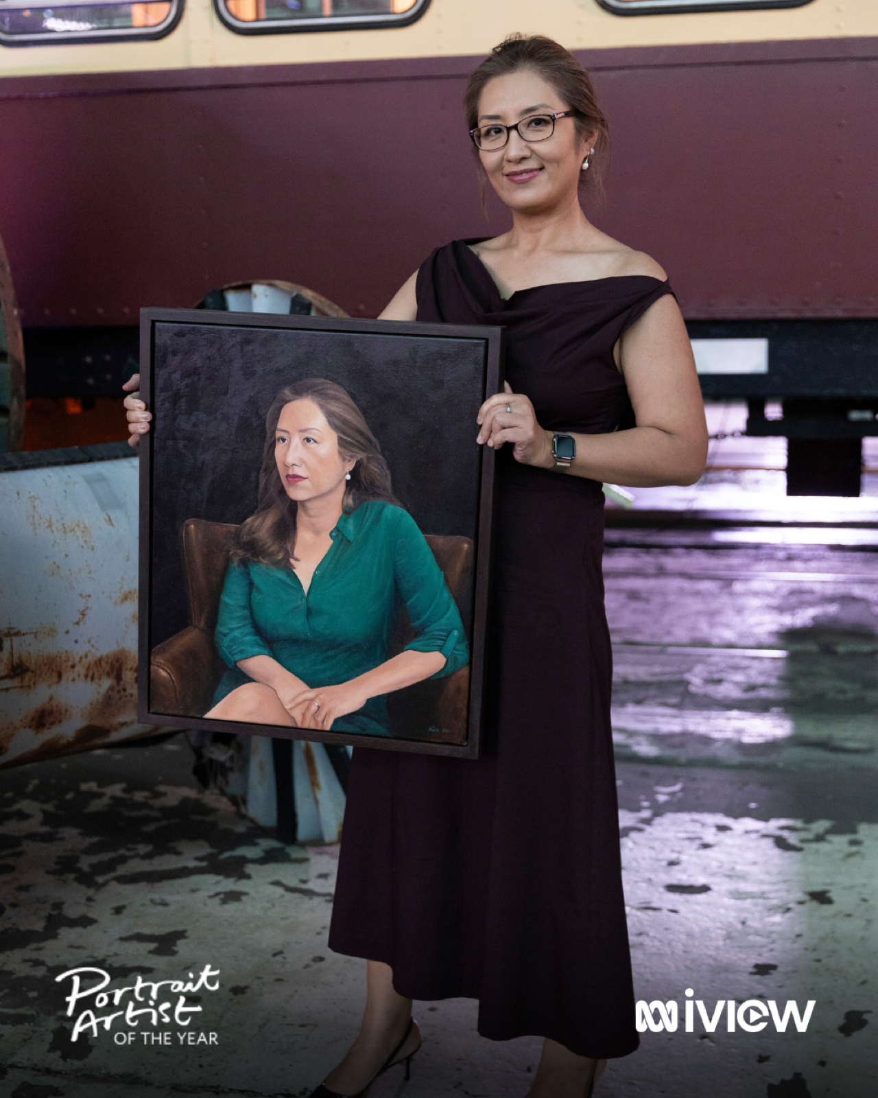 A woman Artist MINA LEE in a brown Witchery dress, wearing glasses and earrings, is holding a framed portrait of another woman. She is standing inside a rustic, partially open building near water.