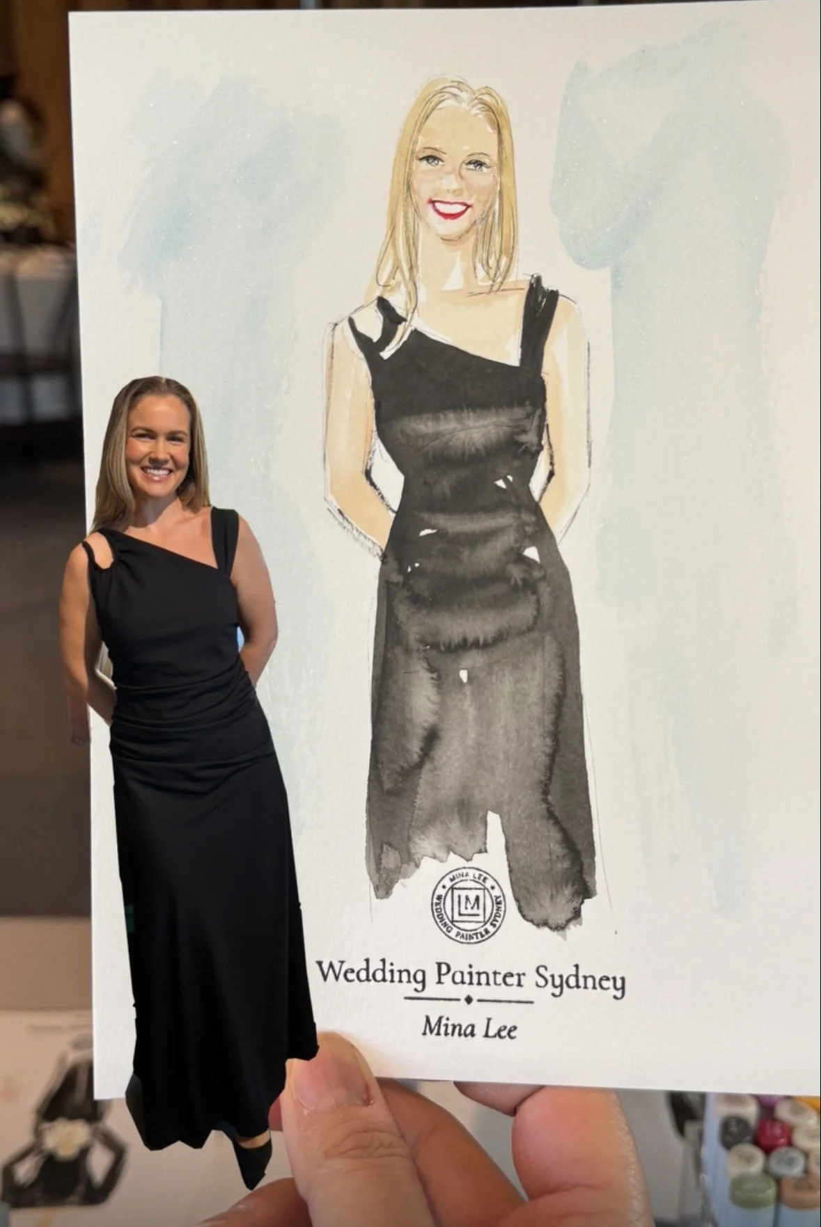 Photograph of a woman standing in front of a large artwork that features a painted portrait of her wearing a black dress. The woman in the photograph is smiling with her hands behind her back, and she is wearing a black dress with a bow on the should