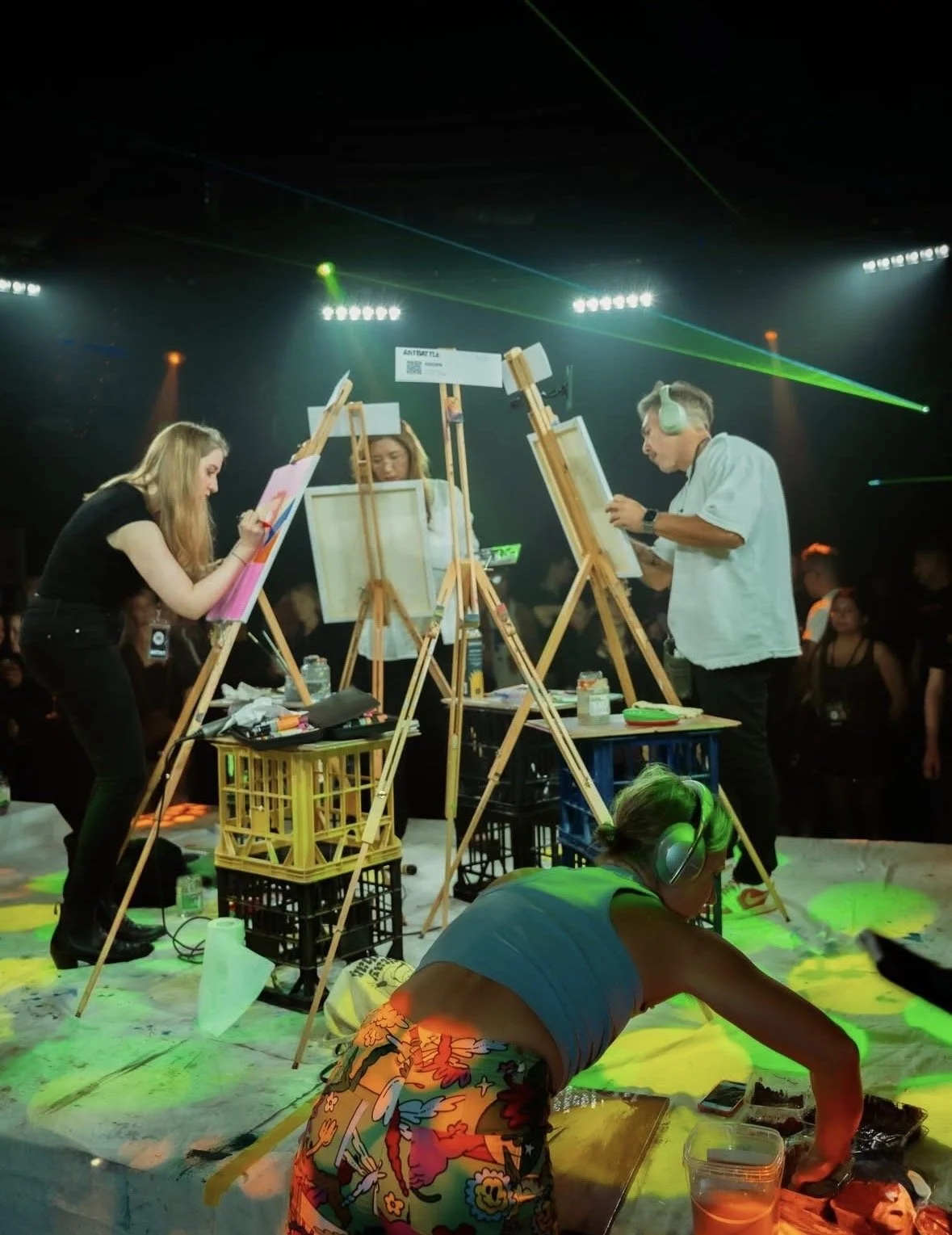 People painting on canvases at an art event with colorful lights, a woman in a gray shirt with headsets working on the foreground, and a lively crowd in the background.