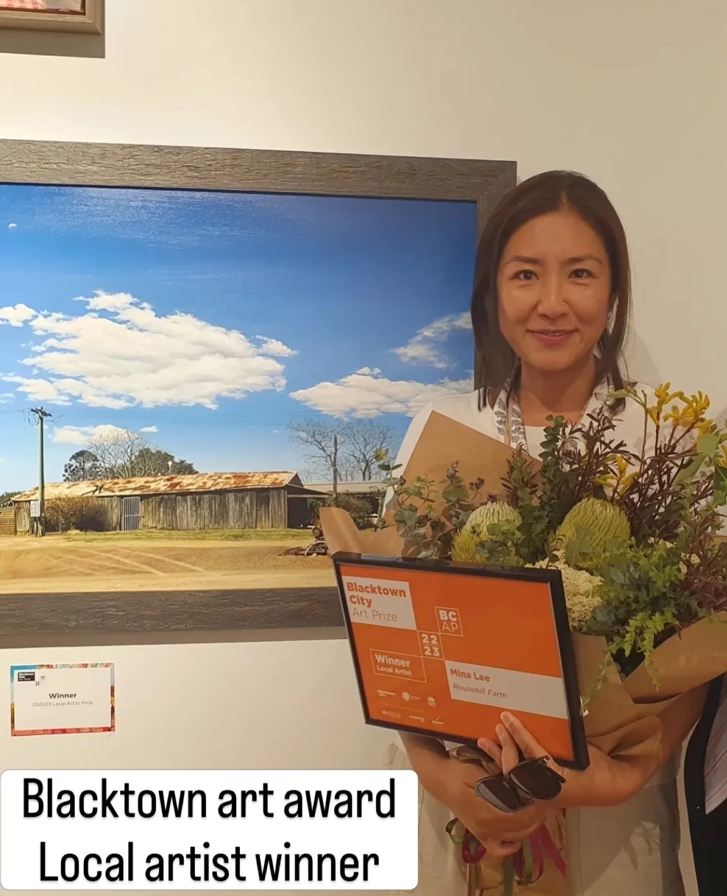 A woman holding a bouquet of flowers and a certificate, standing in front of a landscape painting of a blue sky with clouds and an old barn. The certificate reads "Blacktown art award, local artist winner, Mina Lee, Rousehill Farm."