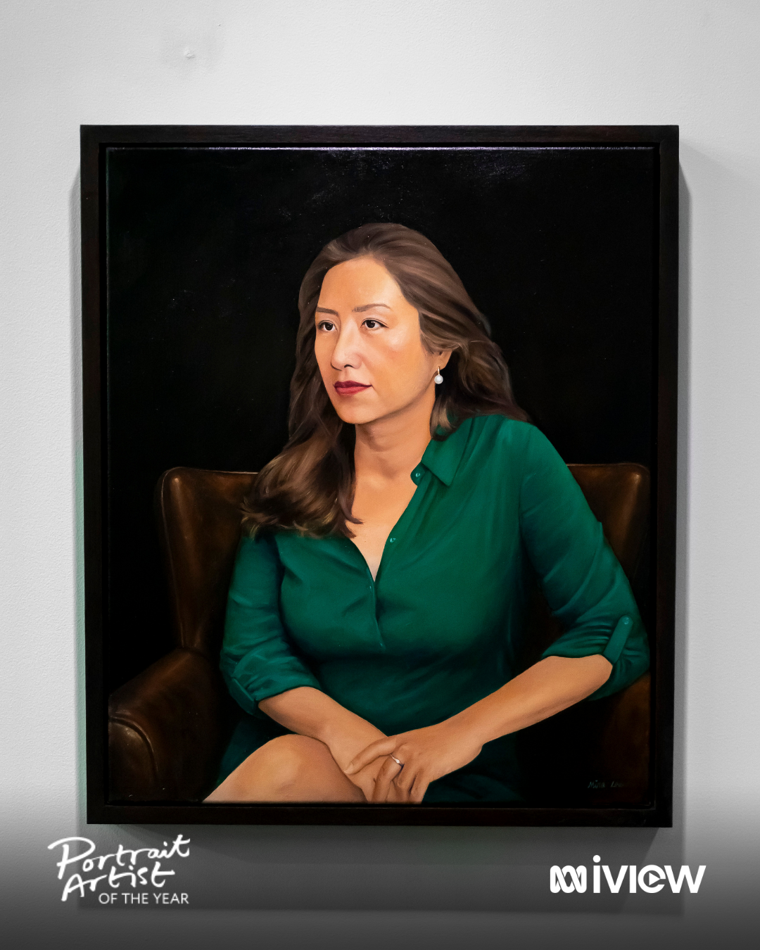 A realistic portrait painting of a woman with medium-length brown hair, wearing a green blouse, sitting on a brown leather chair against a dark background, displayed in an art gallery.