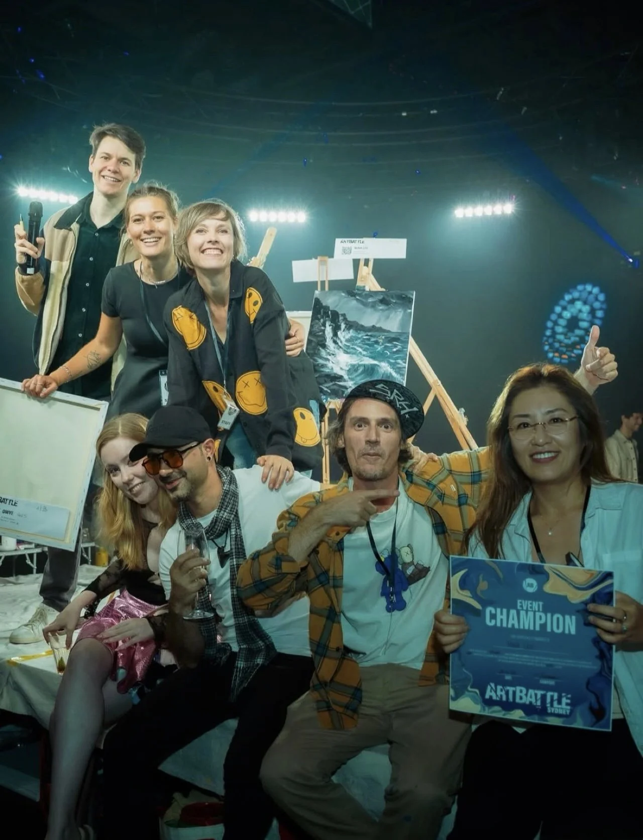 Group of diverse people celebrating at an event, some holding a sign that reads 'Event Champion' and a plaque, with bright stage lights and artwork in the background.