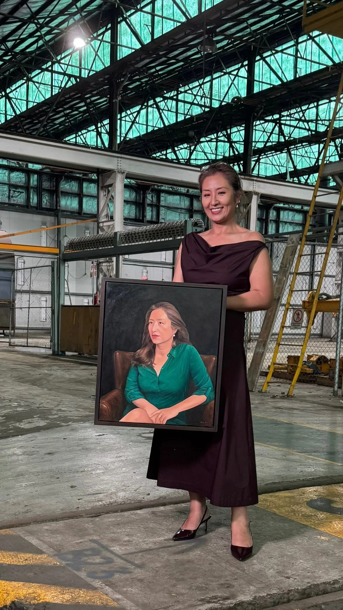 A woman in a dark purple dress and black high heels is holding a framed portrait of a woman wearing a green blouse, inside a large industrial space with metal beams and a high ceiling.