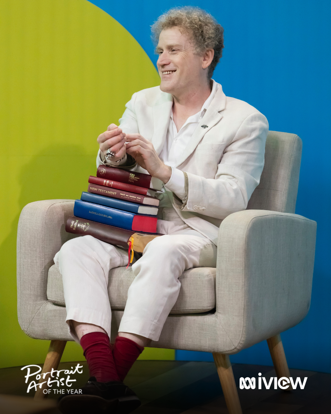 A man in a white suit sitting on a beige armchair holding a stack of books balanced on his lap, smiling and clasping his hands. The background features vibrant blue and green colors with logos for Portrait Artist of the Year and ABC iView.
