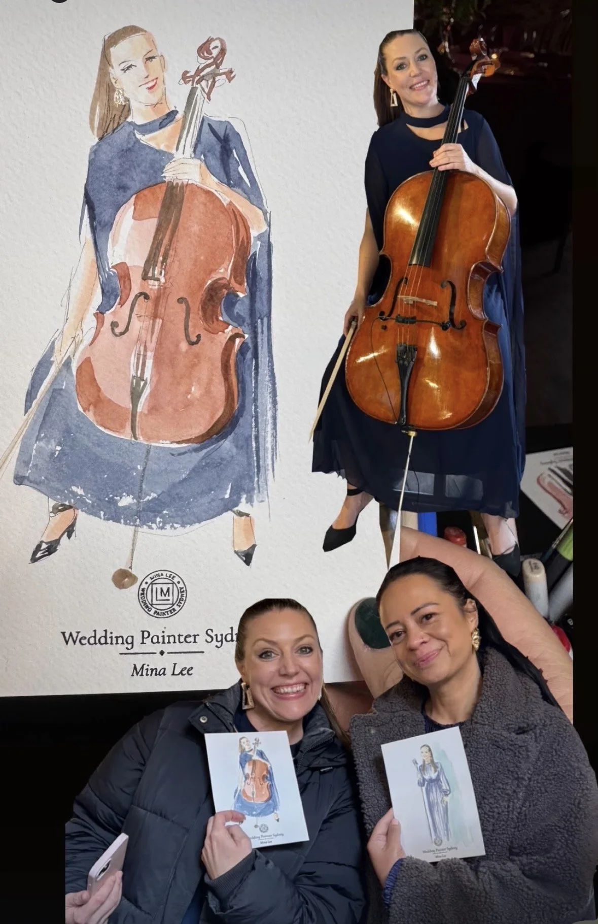 Photo of two women smiling, holding up illustrations of a woman playing a cello, with a poster of a woman holding a cello in the background.