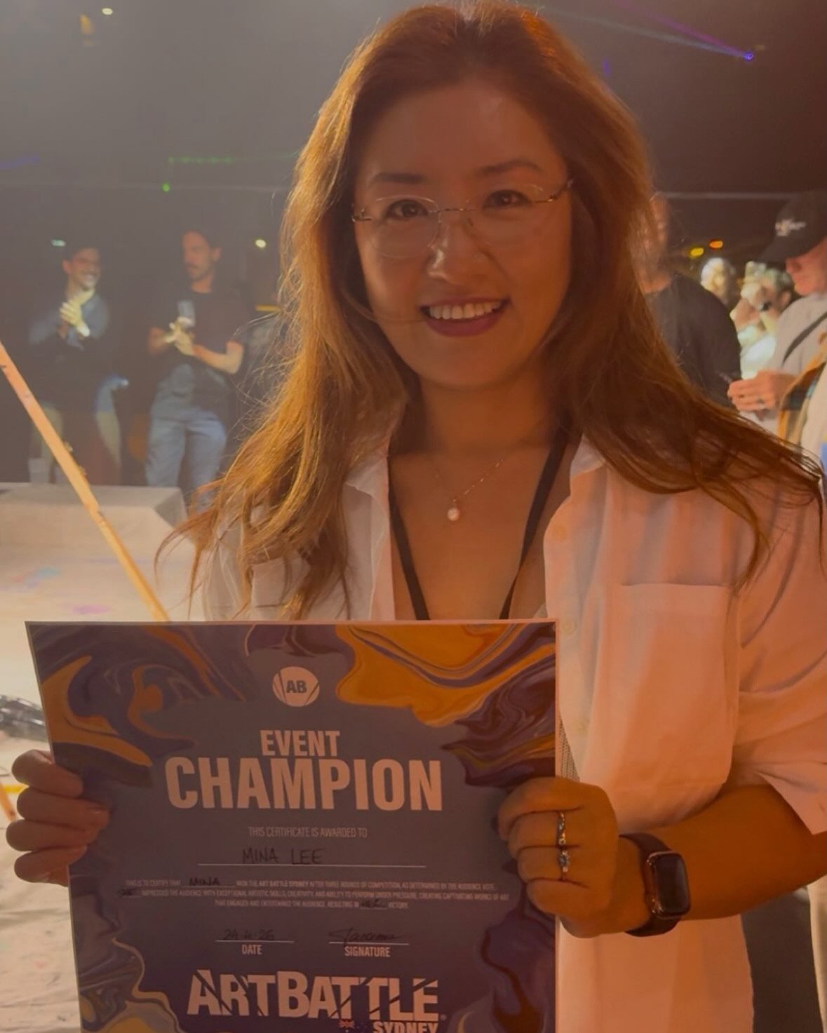 A woman smiling and holding a certificate that reads 'Event Champion' at an event, with people in the background.