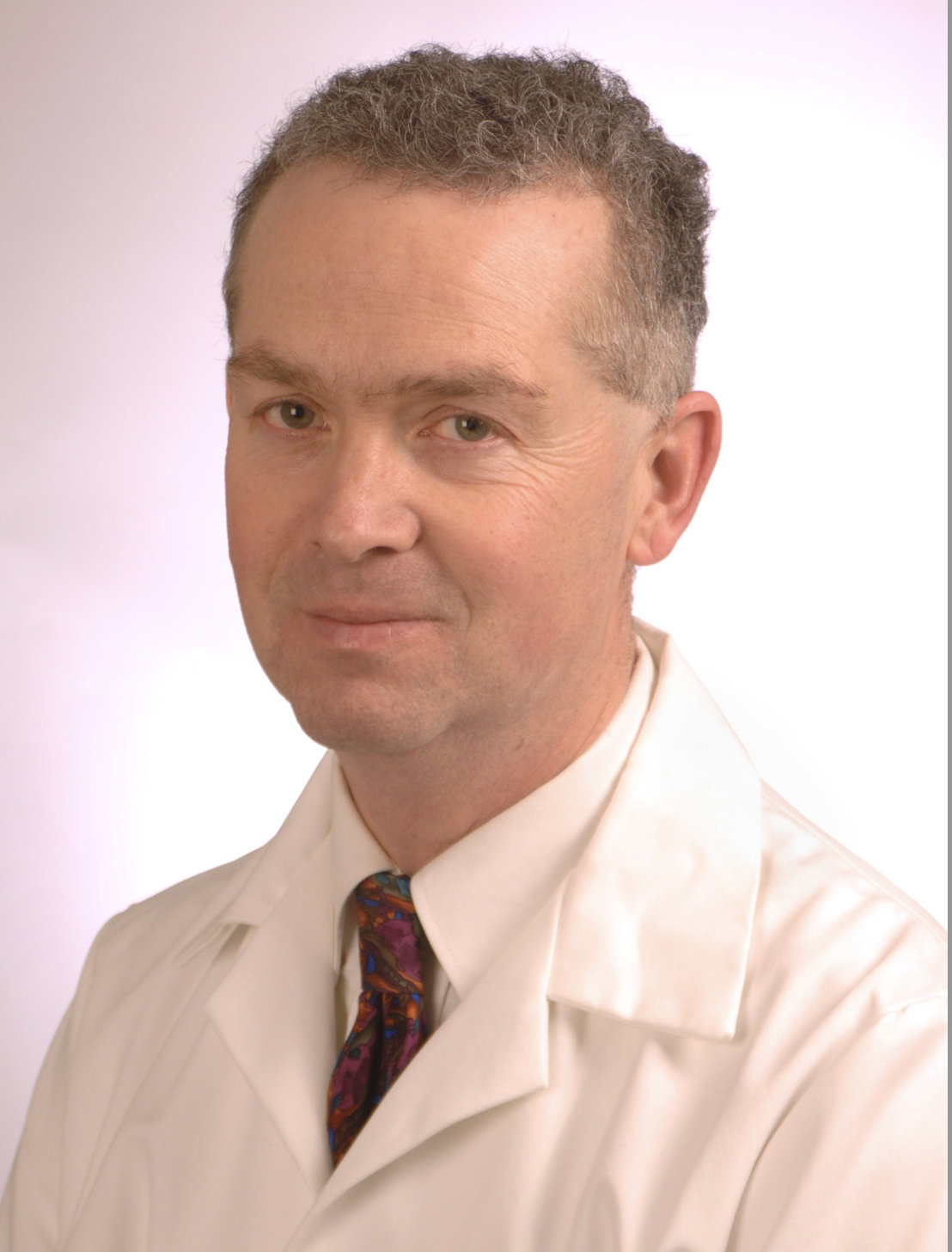 A middle-aged man with short, curly hair wearing a white dress shirt and a colorful necktie, against a plain, light background.