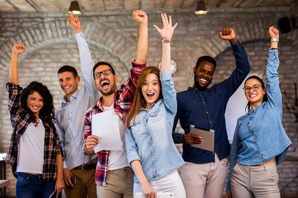 A diverse group of six young adults celebrating indoors with raised fists, smiling and joyful.