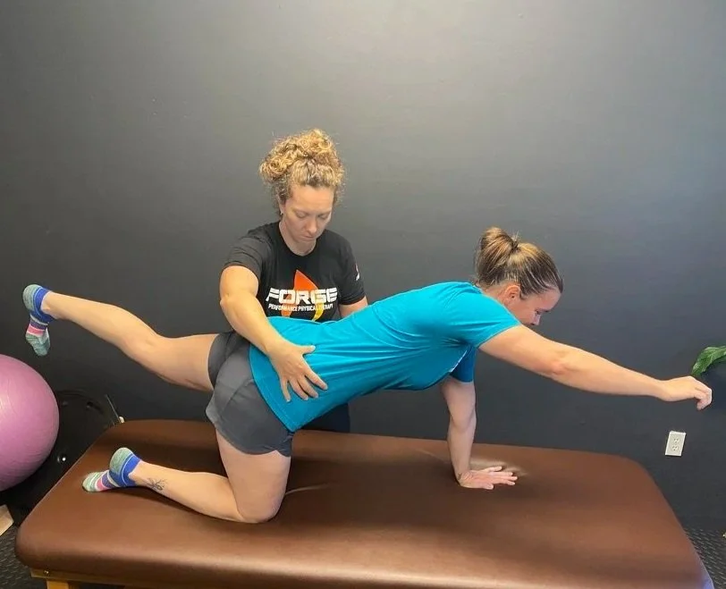 A woman in a blue shirt and gray shorts performs a physical therapy exercise on a padded table with assistance from a trainer.