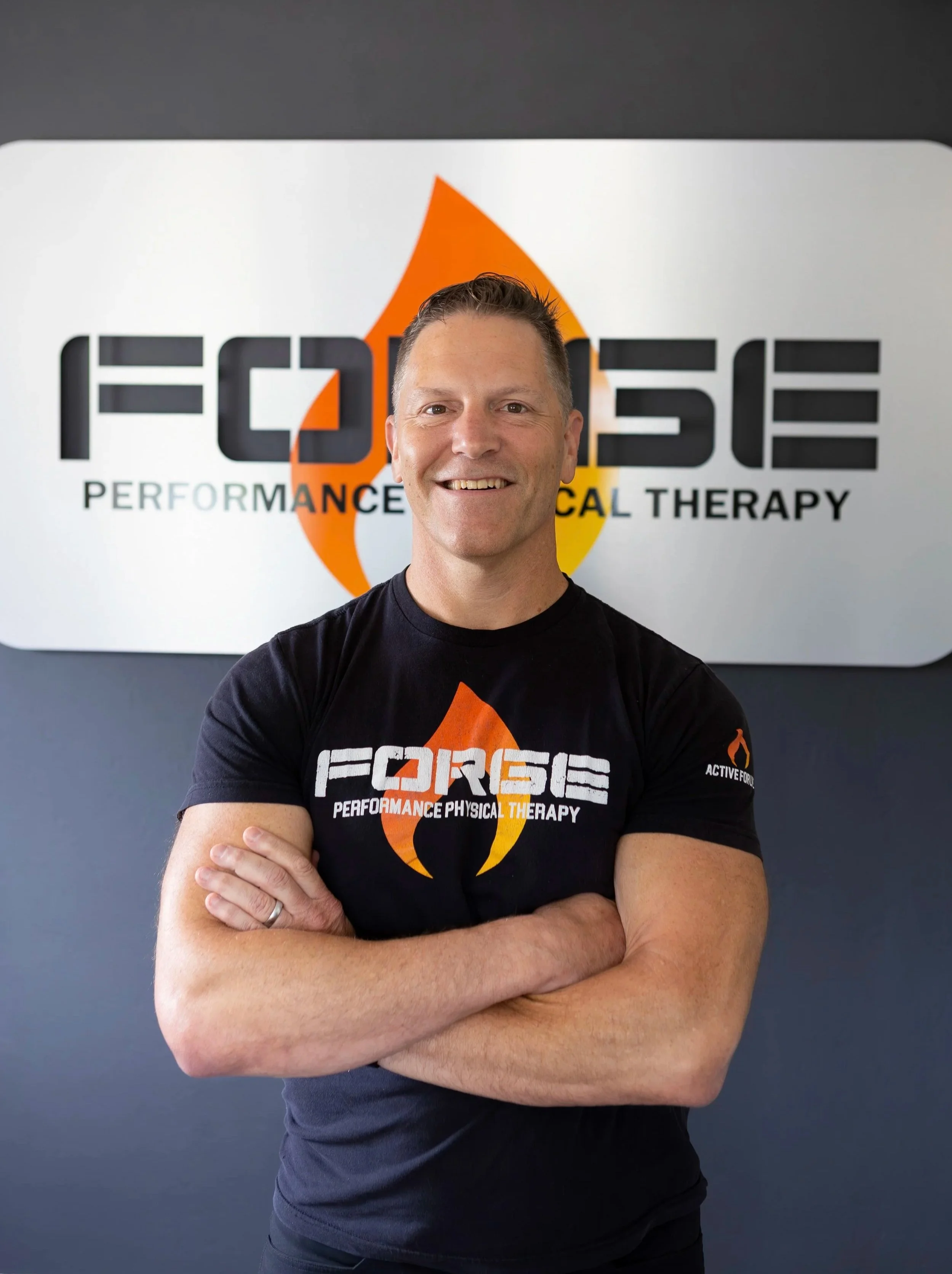 A man with short hair and a smile stands with arms crossed in front of a sign that reads 'FORGE Performance Physical Therapy' with a flame logo.