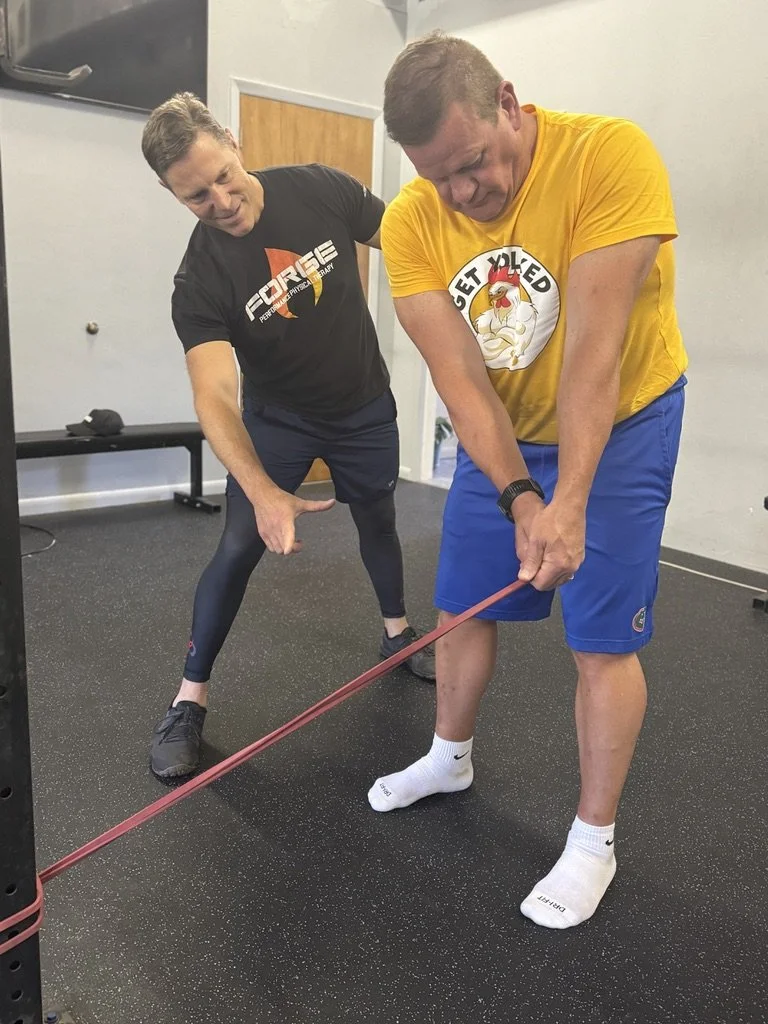 A fitness trainer assisting a man with a resistance band exercise in a gym. The man is wearing a yellow T-shirt with a chicken logo and blue shorts, while the trainer is in a black T-shirt with gym branding and black leggings.