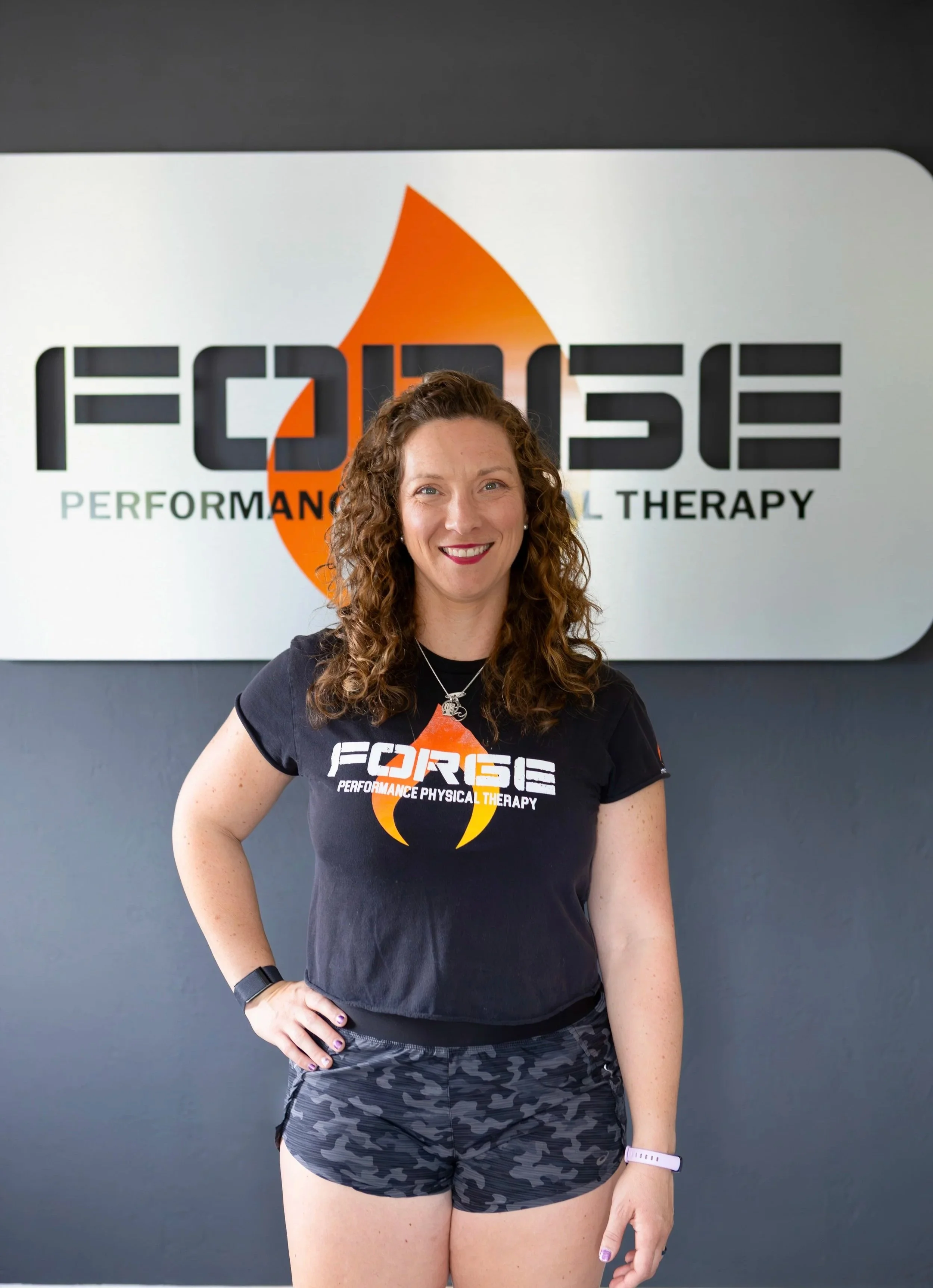 A woman with curly hair smiling, wearing a black T-shirt with the logo and text of Forge Performance Physical Therapy, standing in front of a large Forge Performance Physical Therapy sign.