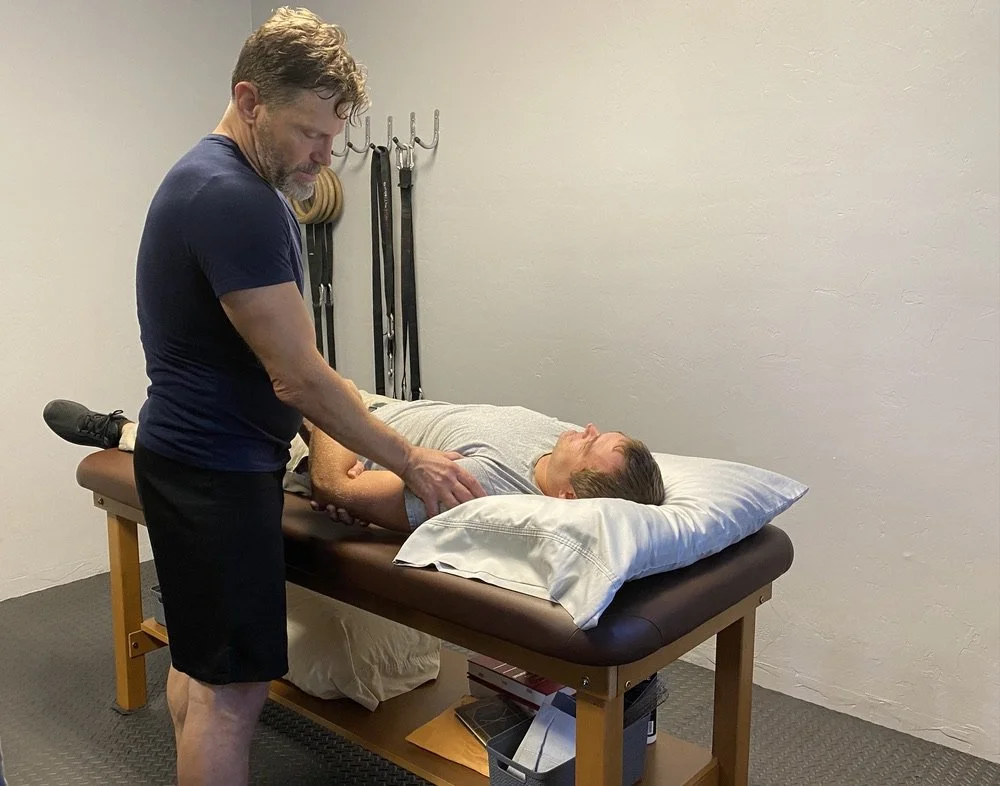 A man lying on his back on a massage table with a pillow under his head, receiving gentle therapy from a therapist who is standing beside him, in a room with exercise equipment hanging on the wall.