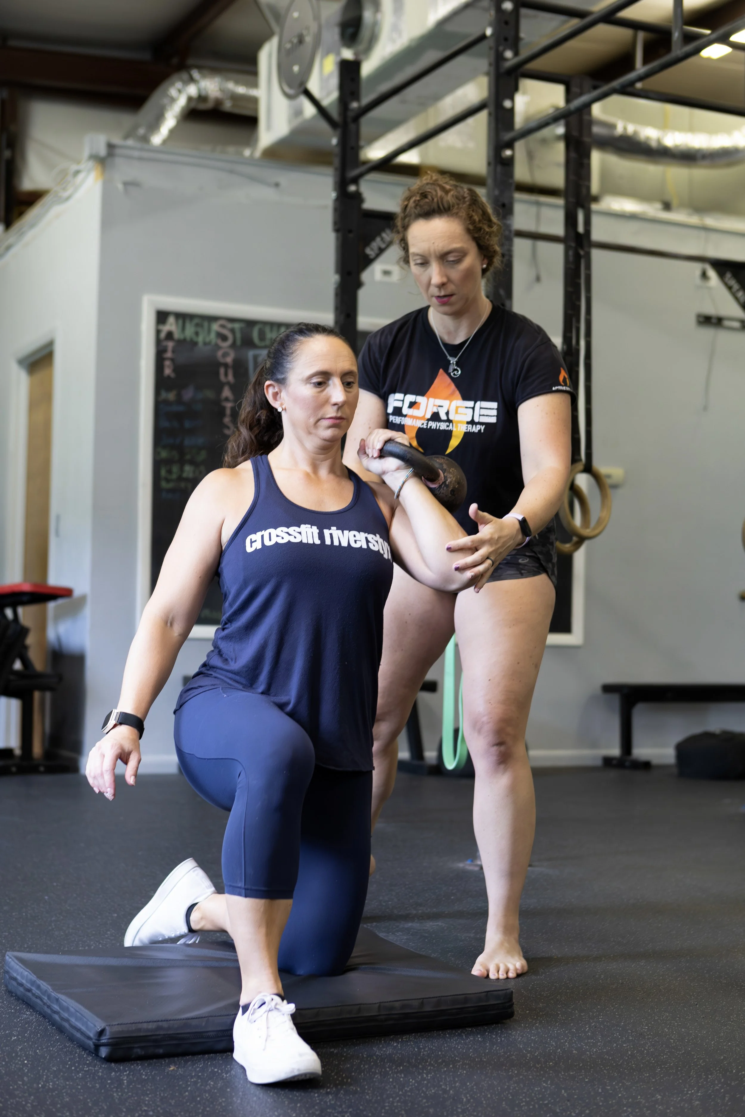 A woman wearing a navy tank top with 'crossfit inverso' and navy leggings kneels on a cushioned mat while gripping a kettlebell held by a trainer in a gym. The trainer, in a black T-shirt with orange and white logo, assists her with the exercise.