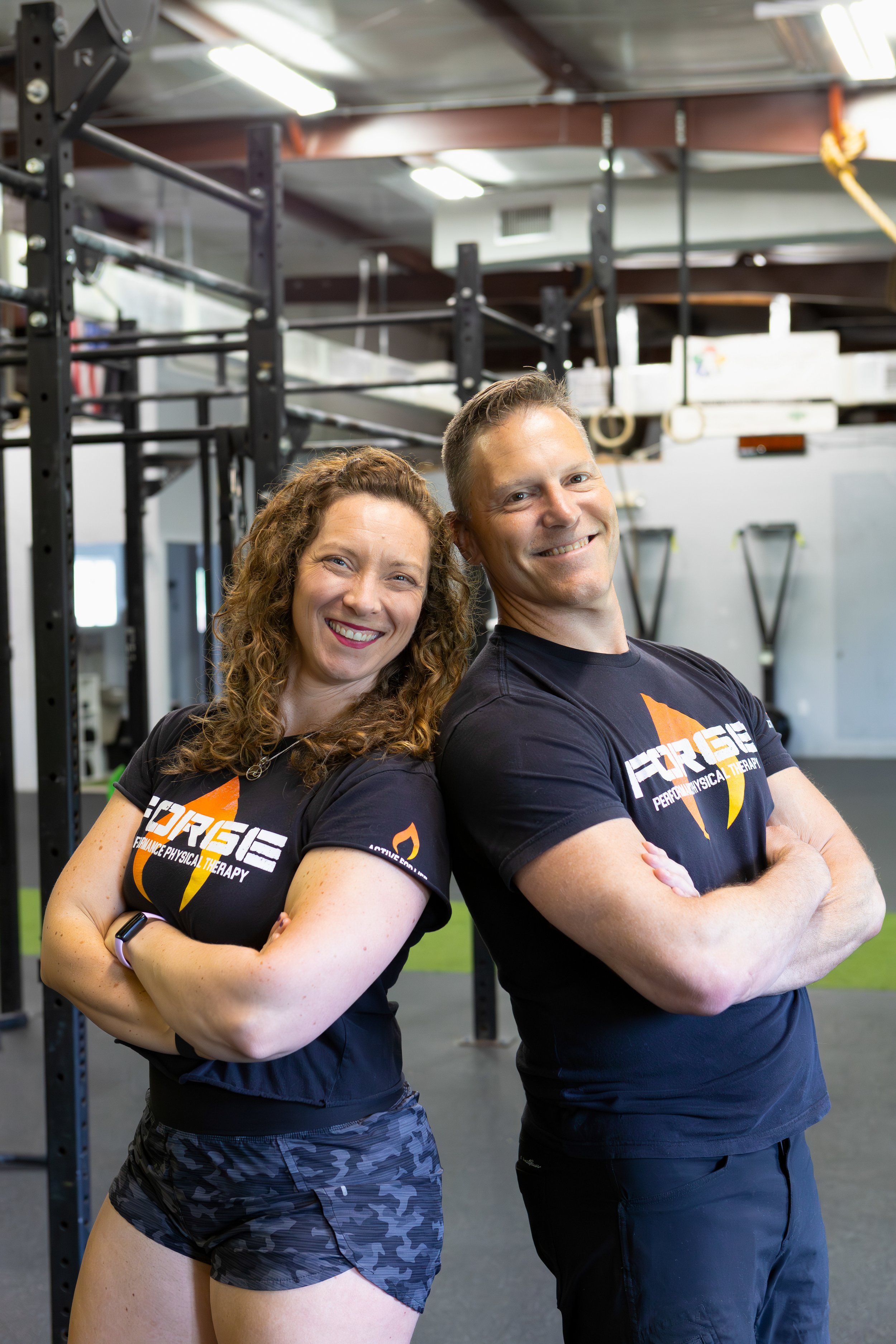 Two smiling fitness trainers standing back to back with arms crossed in a gym, wearing black shirts with a logo that says 'FORCE' and 'Performance Physical Therapy,' with gym equipment and rings visible in the background.