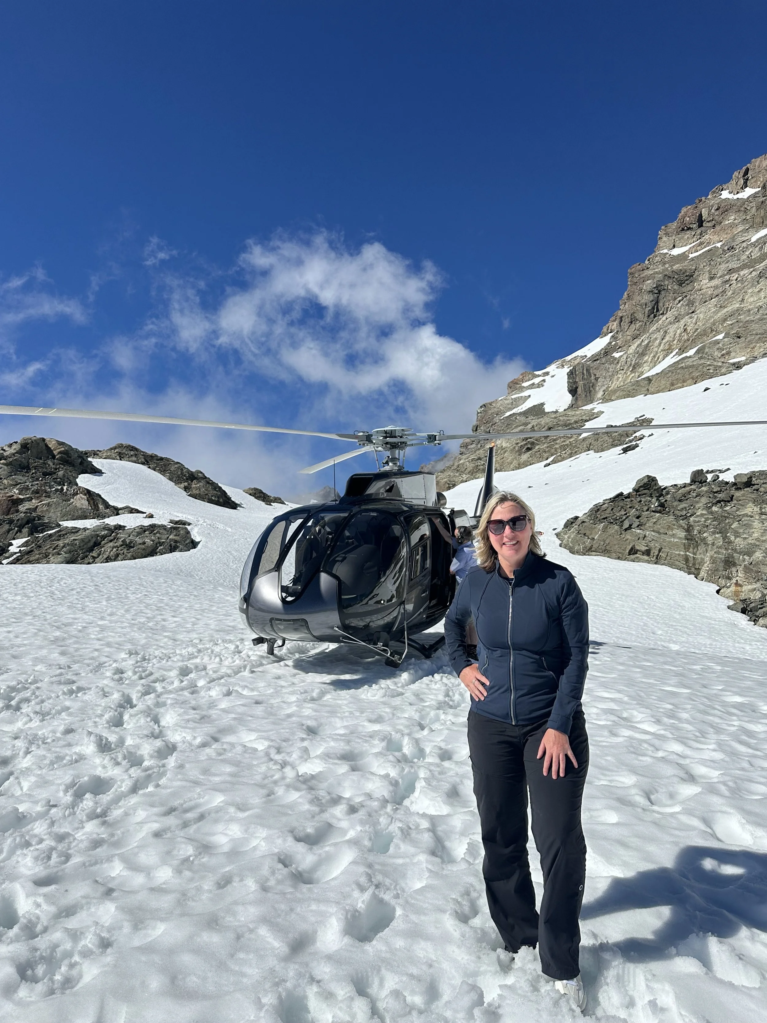 A woman in sunglasses and outdoor gear standing on snowy mountain terrain in front of a black helicopter with a rocky mountain and blue sky in the background.