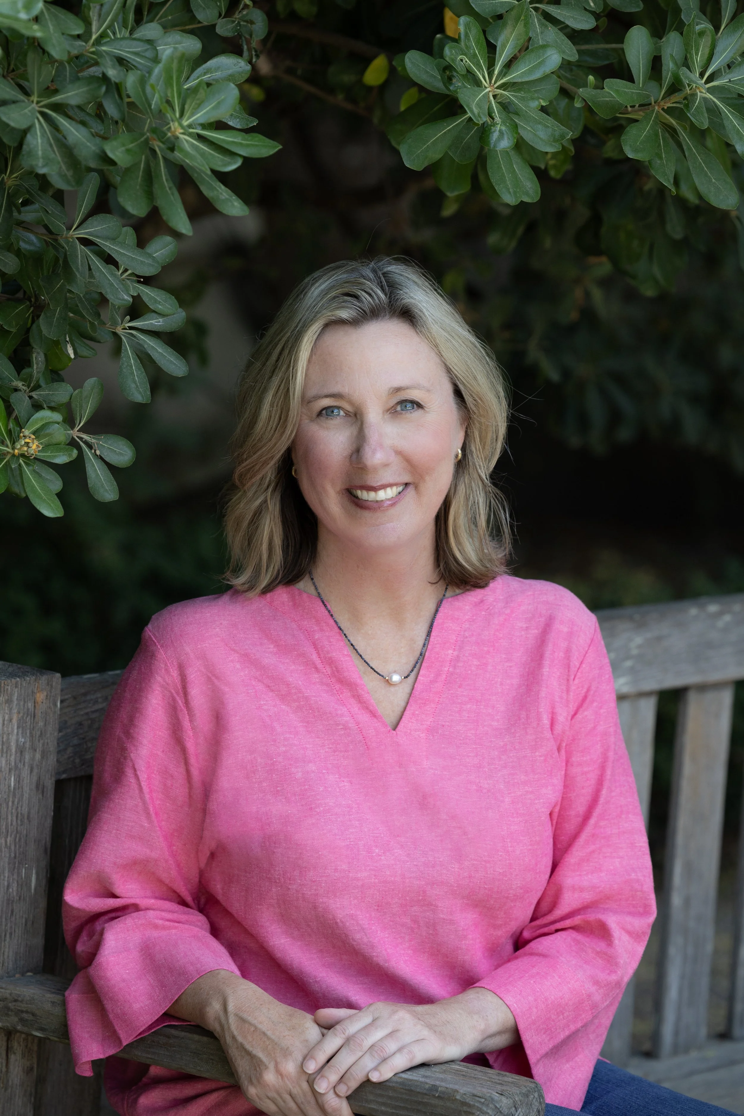 A woman with blonde hair and blue eyes sitting outdoors on a wooden bench, wearing a pink top and a necklace with a pearl, smiling at the camera with green foliage in the background.