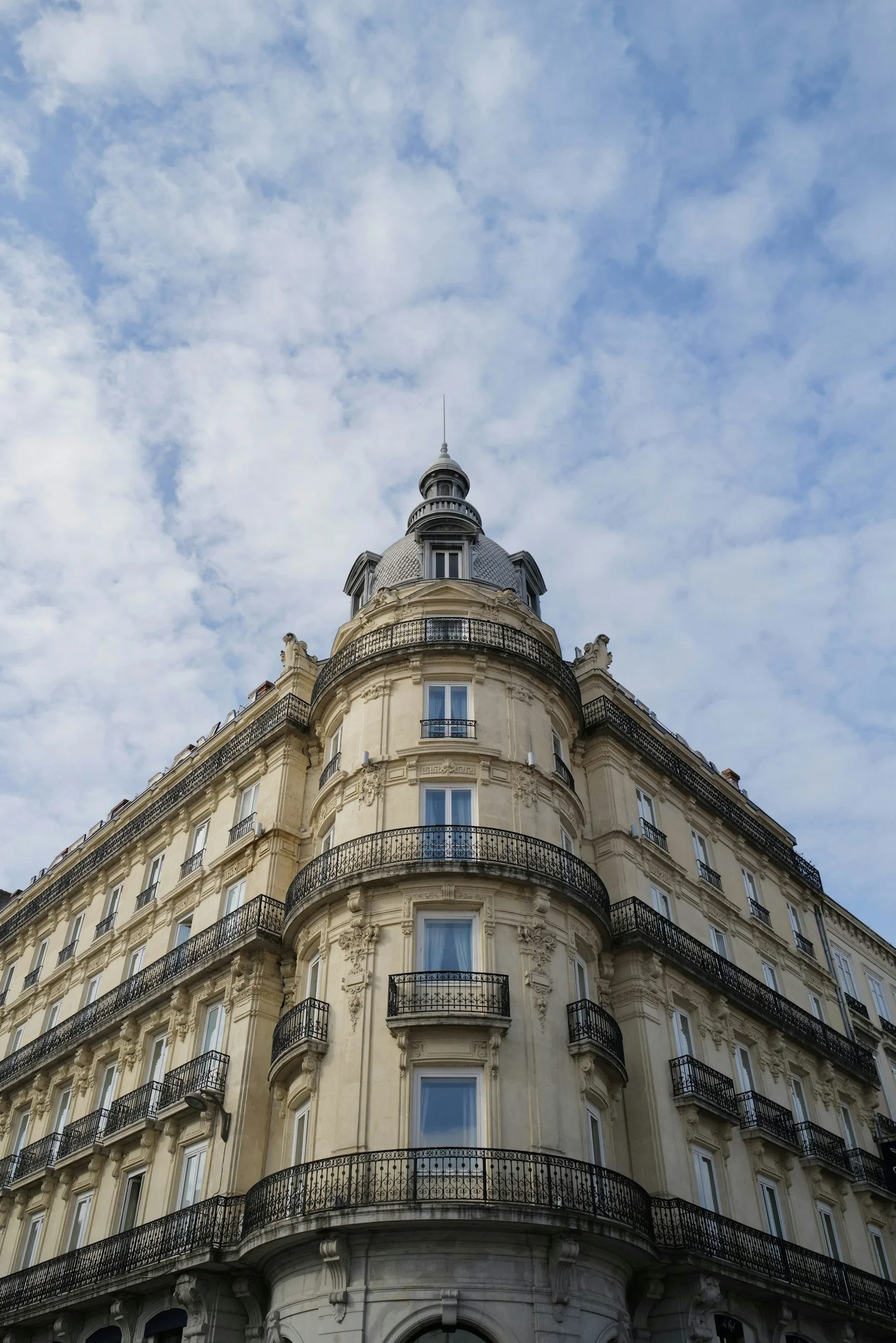 A tall, ornate, multi-story building with balconies and a rounded tower at the corner, topped with a dome and spire, against a blue sky with scattered clouds.