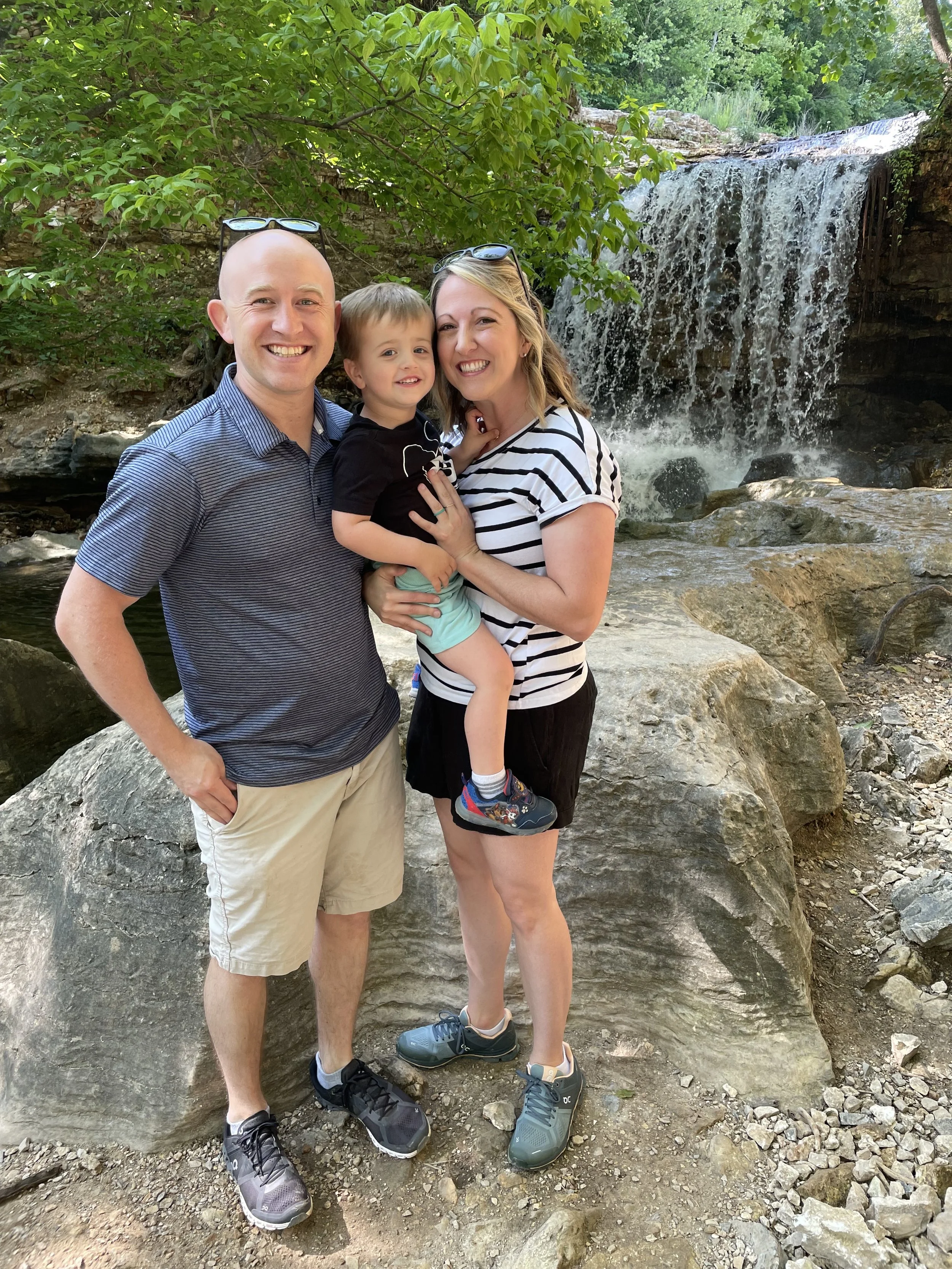 Family of three standing on rocks near a waterfall surrounded by green trees during daytime