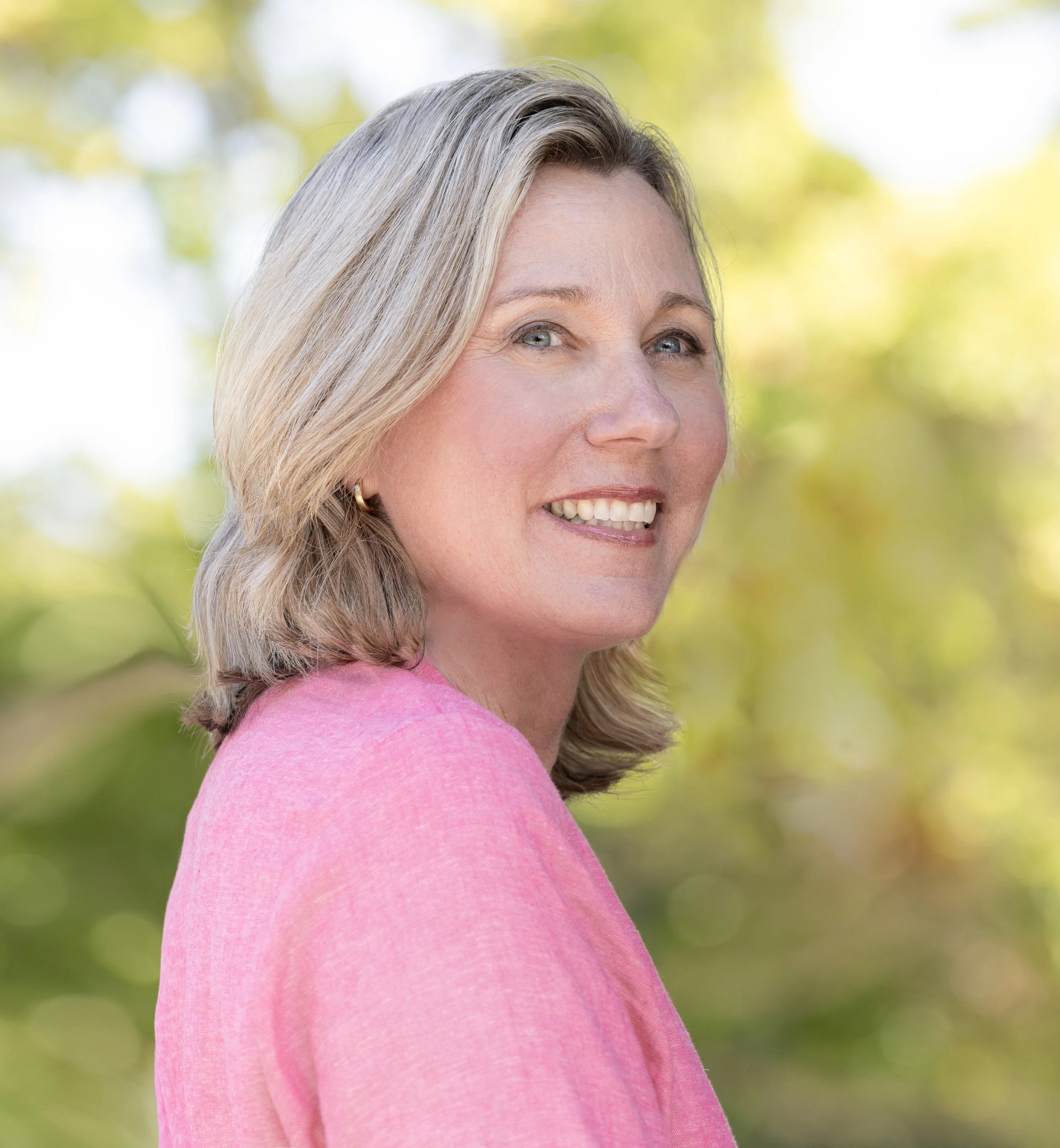 A woman with shoulder-length blonde hair smiling outdoors, wearing a pink top, with blurred green trees in the background.