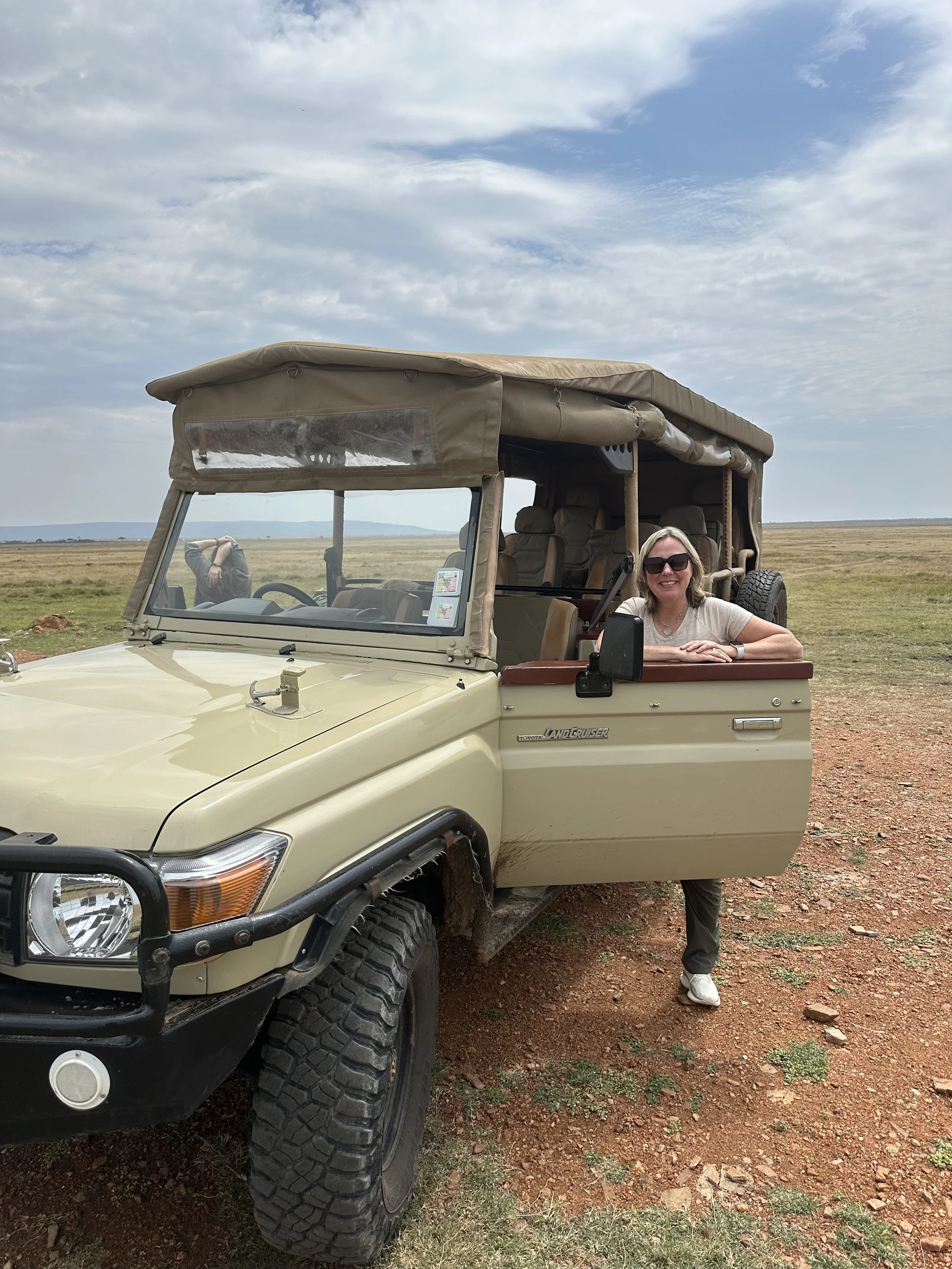 Woman smiling and wearing sunglasses leaning on an open door of a beige Toyota Land Cruiser parked on a grassy plain under a partly cloudy sky.