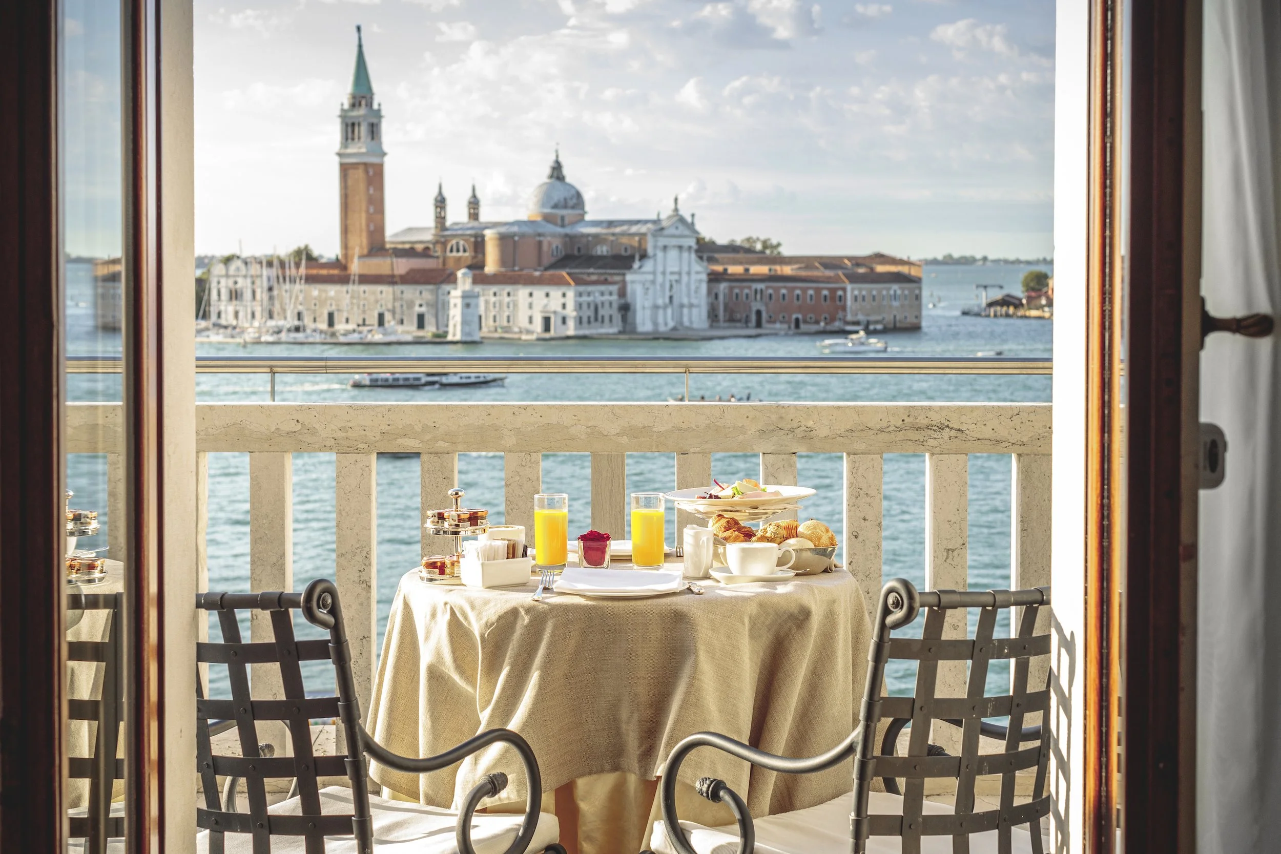 Breakfast table set on a balcony overlooking a waterscape with historic buildings, including a church with a tall bell tower, in the background.