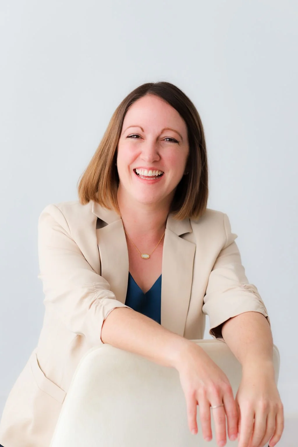 A woman with shoulder-length brown hair smiling, wearing a beige blazer and a blue top, sitting with her arms resting on a white chair against a plain light background.