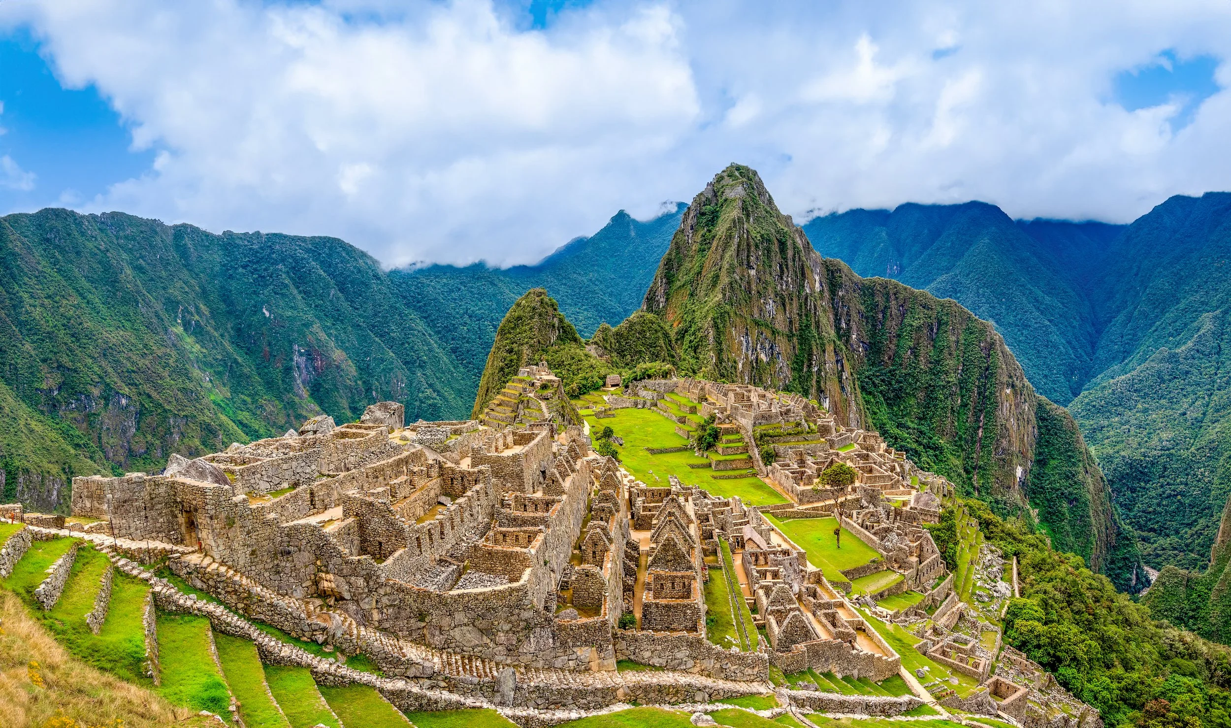 Ancient Incan ruins of Machu Picchu with terraces and stone structures surrounded by green mountains under a cloudy sky.