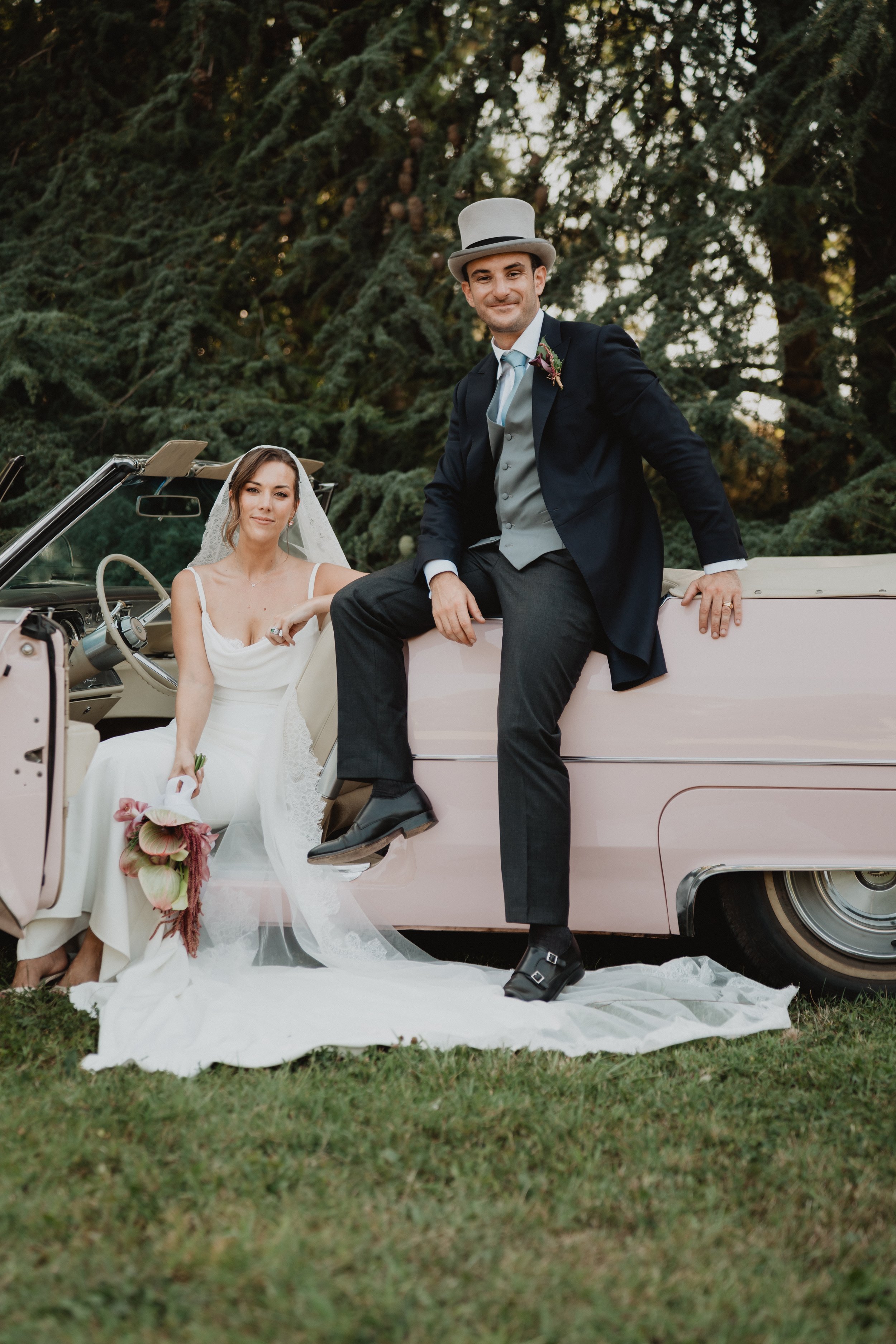 Un couple de mariés posant à côté d'une vieille voiture vintage, la femme en robe de mariée blanche et le homme en costume avec un chapeau haut de forme, dans un espace extérieur avec des arbres en arrière-plan.