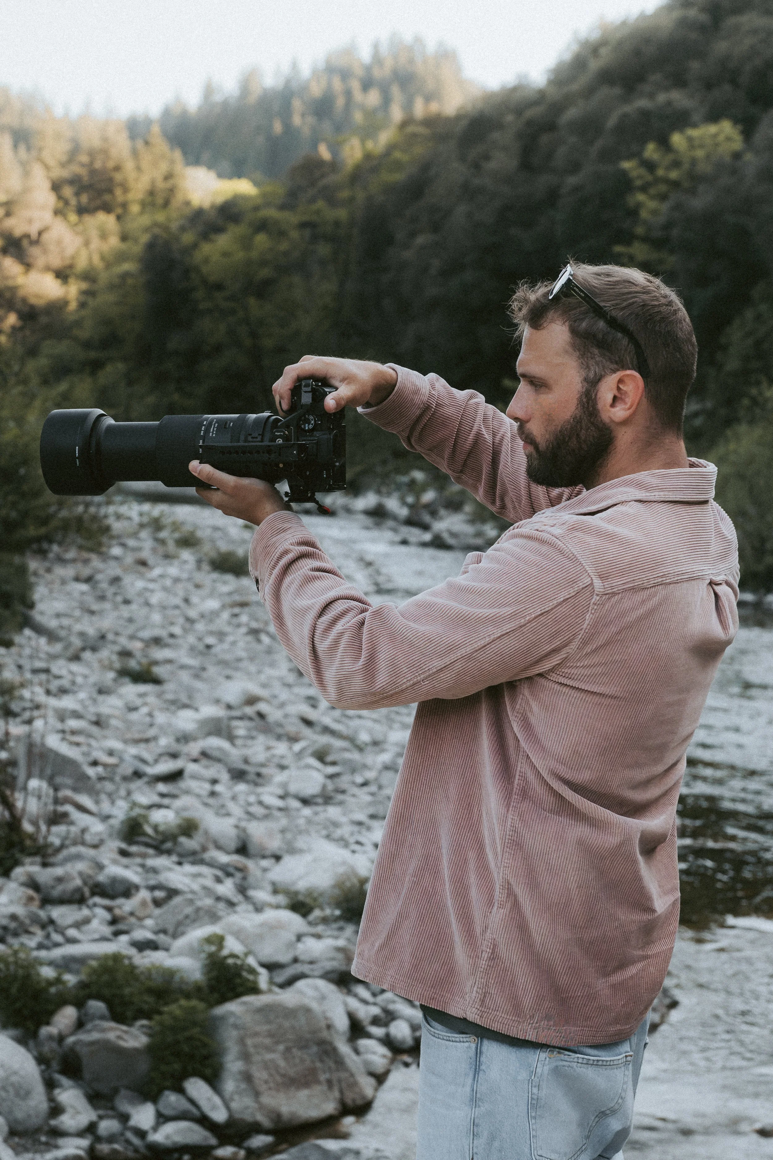 Un homme photographie avec un appareil photo à longue focale à l'extérieur, près d'une rivière et de montagnes verdoyantes, en fin d'après-midi.