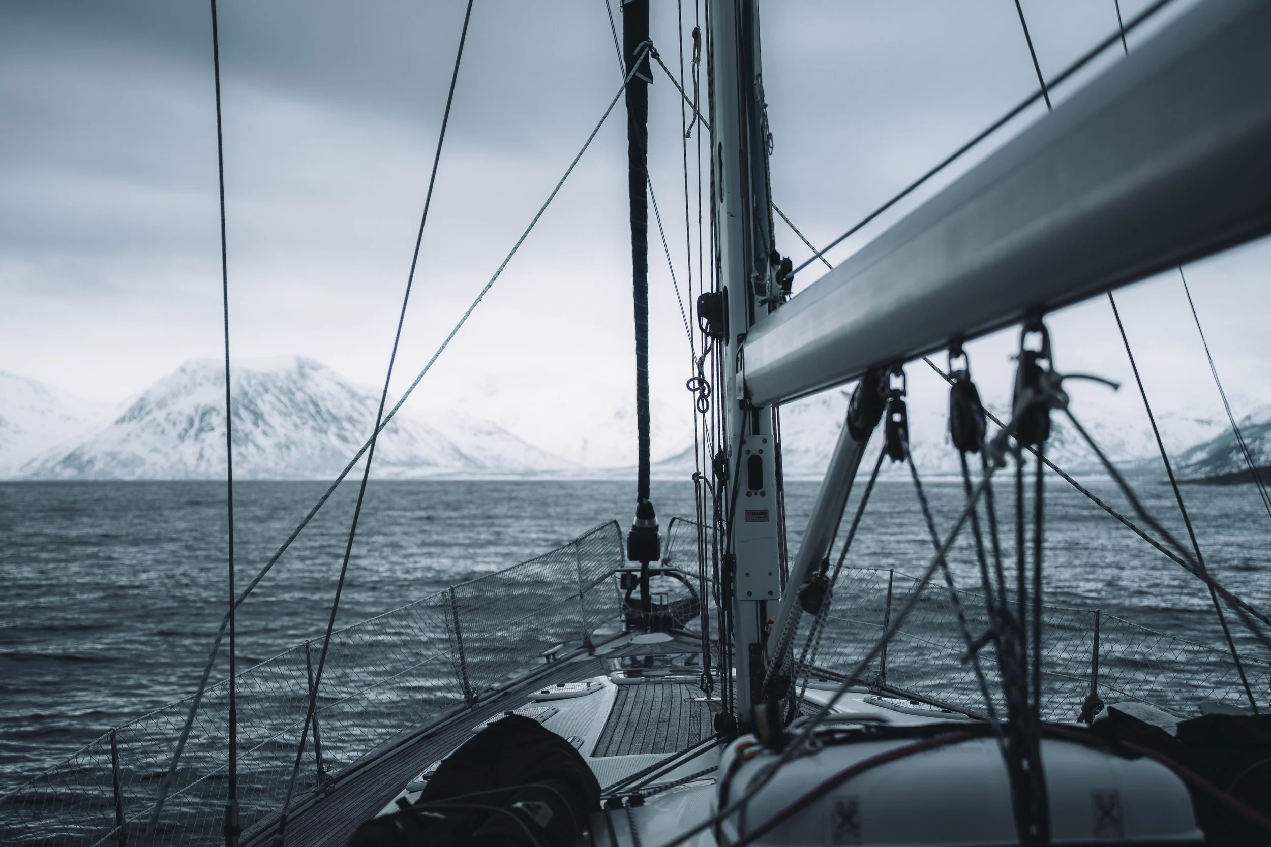 Vue depuis un bateau montrant la proue avec les cordages, voiles et le mât, sur un lac ou océan avec des montagnes enneigées en arrière-plan, ciel nuageux.