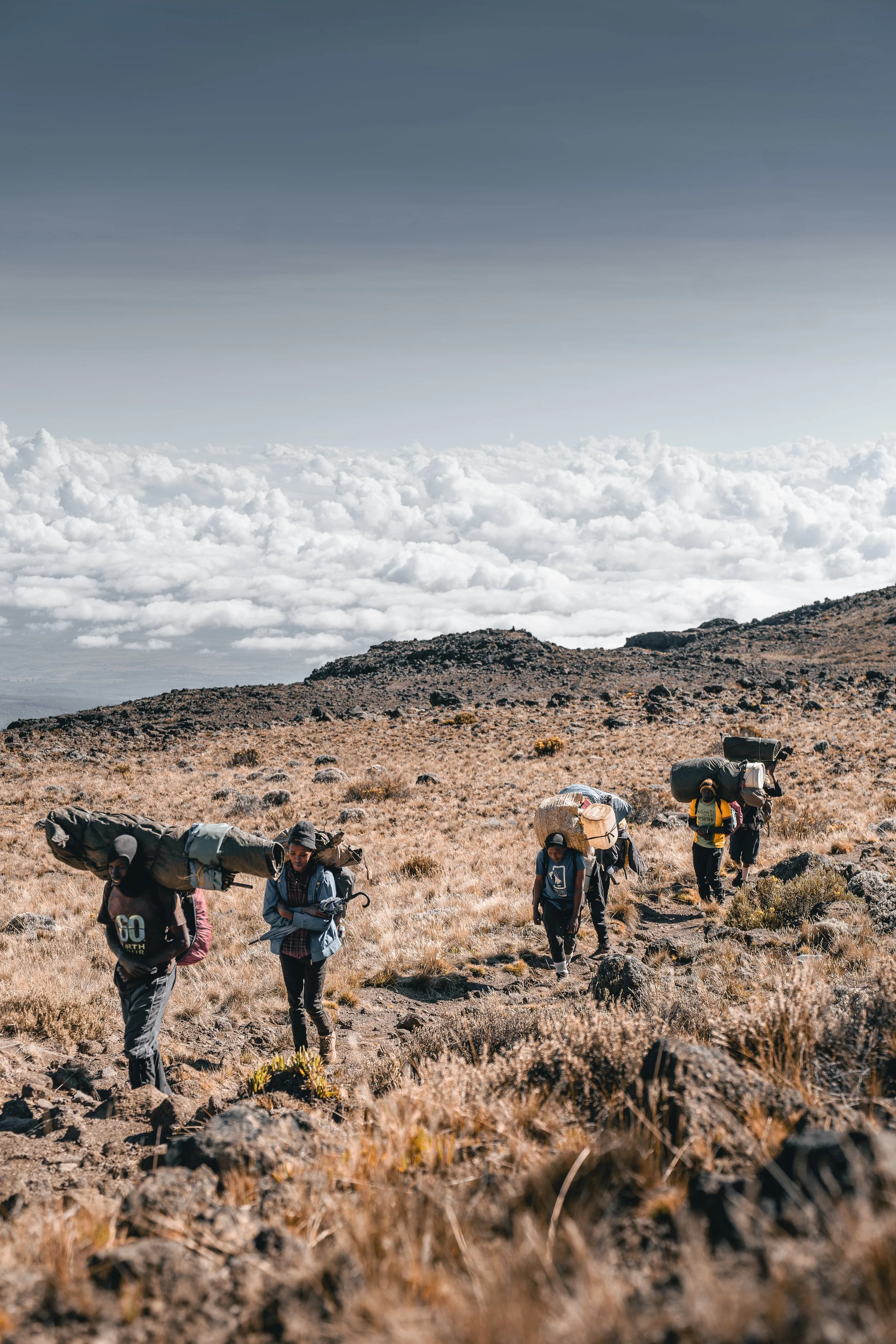 groupe de randonneurs marchant sur un sentier rocheux en montagne, portant de gros sacs à dos, sous un ciel nuageux.