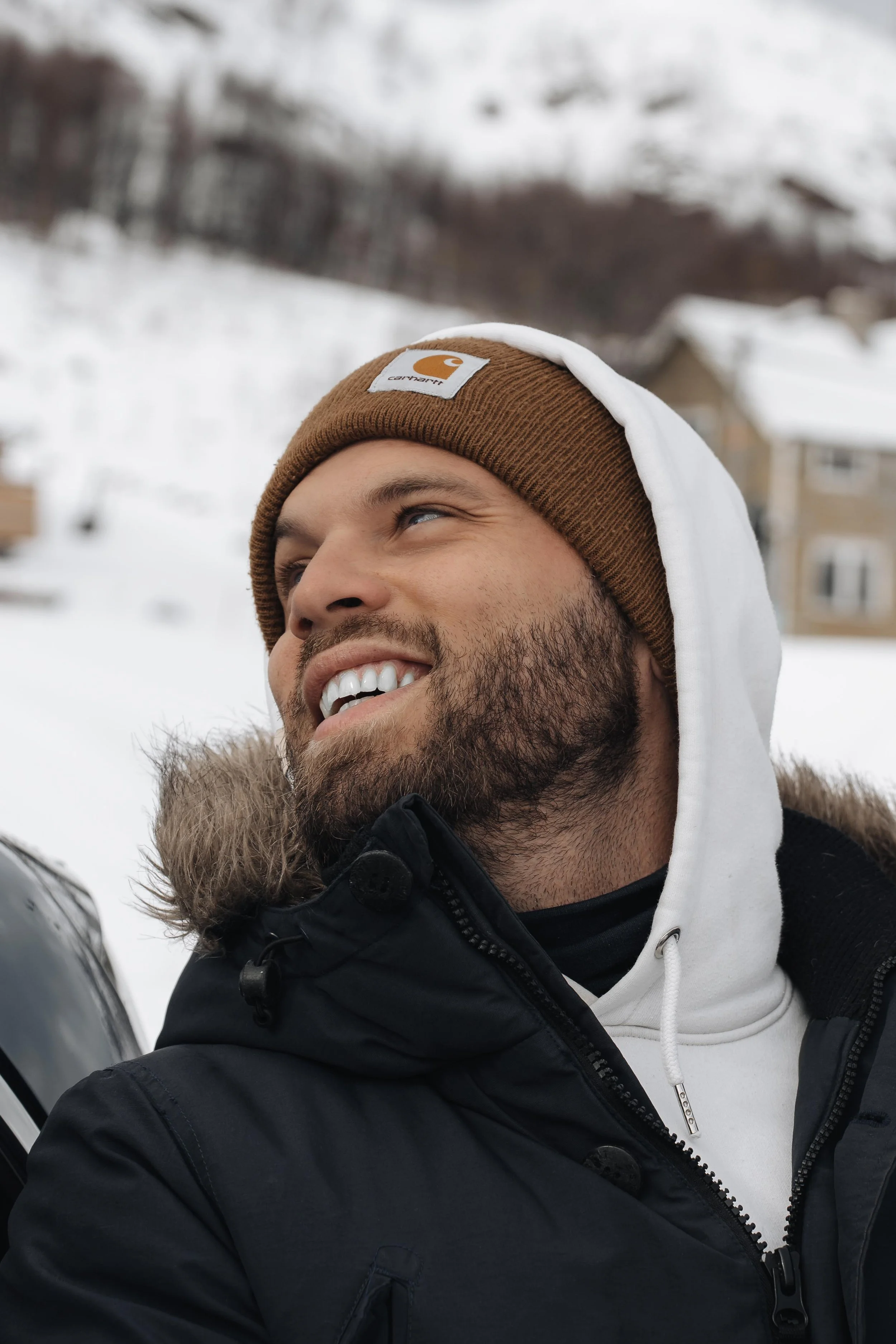 Un homme souriant, portant un bonnet marron avec un logo Carhartt, une veste noire avec une capuche en fourrure et un sweat à capuche blanc dans un environnement enneigé.