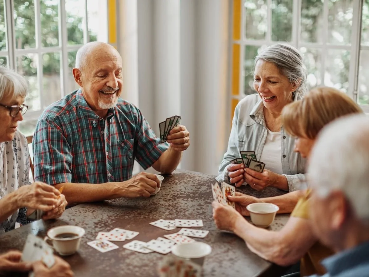 Five elderly people playing a card game at a table, smiling and laughing, with cups of coffee.