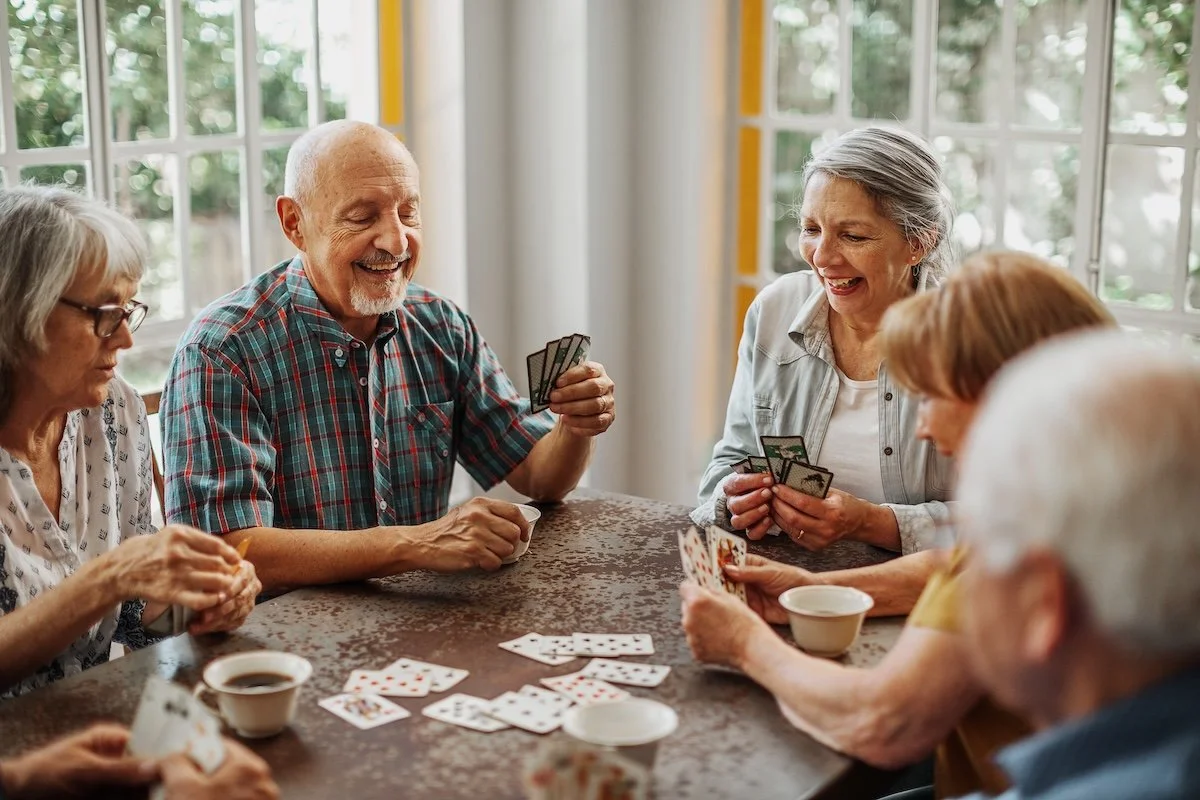 Six elderly people playing cards and laughing together around a wooden table in a bright room with large windows.