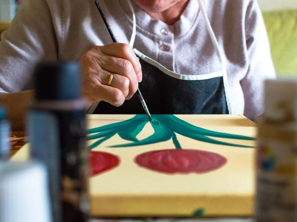 An elderly person painting cherries on a canvas with a small paintbrush. The person is wearing a gray shirt and a black apron, and is focused on their artwork.