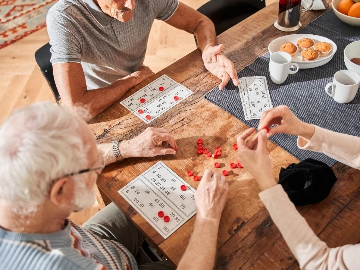 Three elderly women playing bingo at a wooden table with snacks and drinks, using bingo cards and red markers.