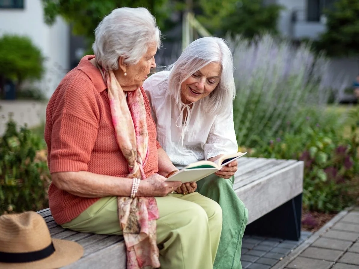 Two elderly women sitting on a park bench, reading books and smiling at each other, with greenery and purple flowers in the background.